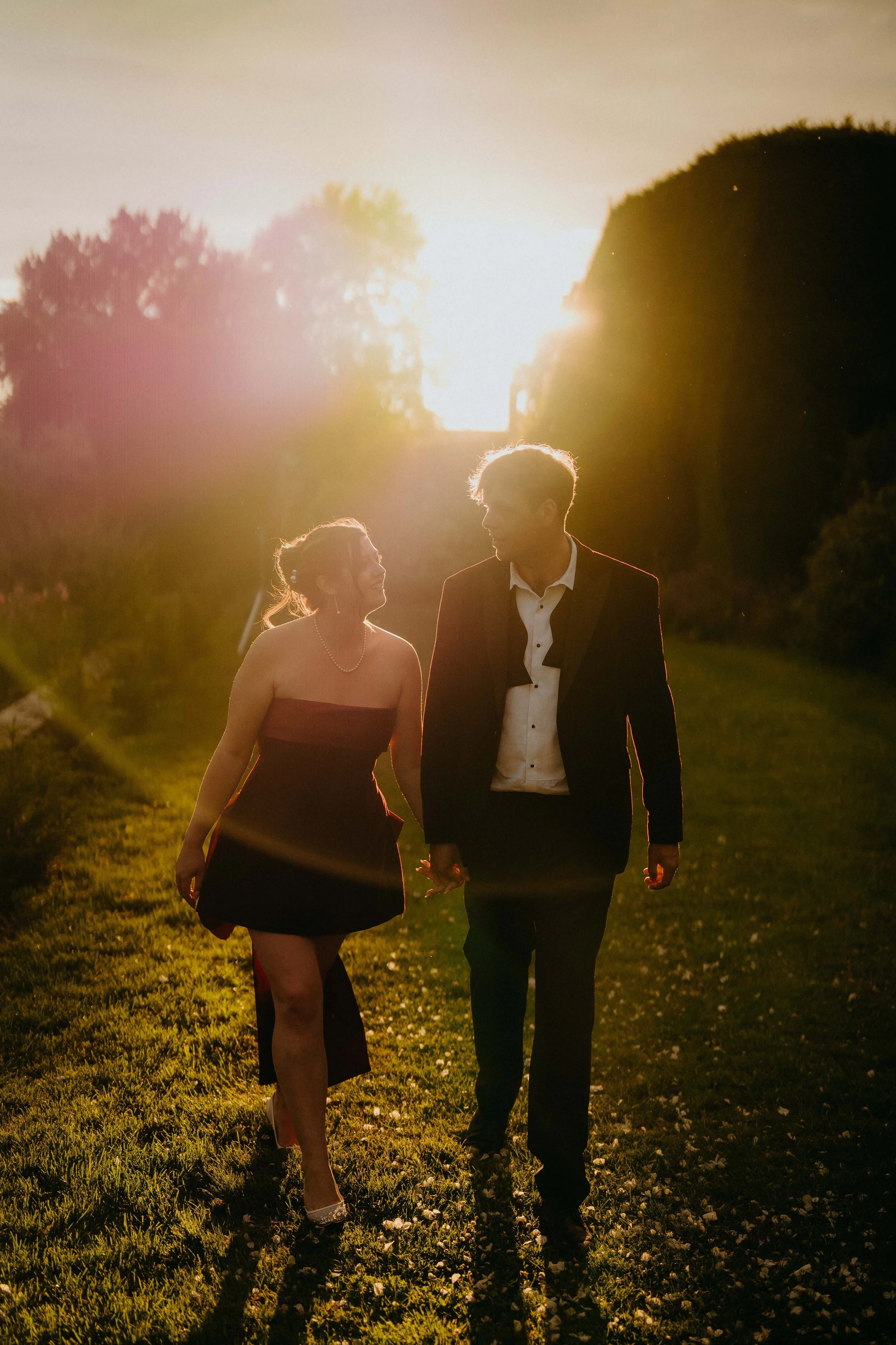 A couple walking hand in hand outdoors during sunset, with trees in the background and grass underfoot, dressed in formal attire.