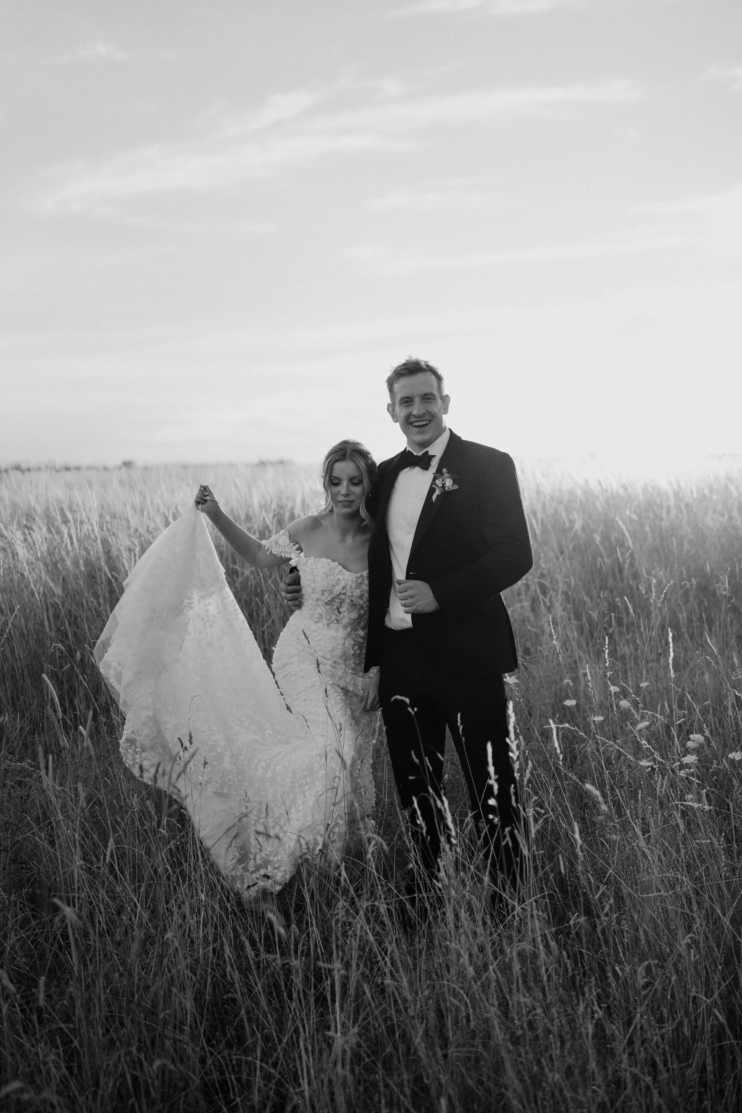 A black-and-white photo of a bride and groom smiling and holding hands in a tall grassy field, with a cloudy sky in the background at Stone Barn in the cotswolds