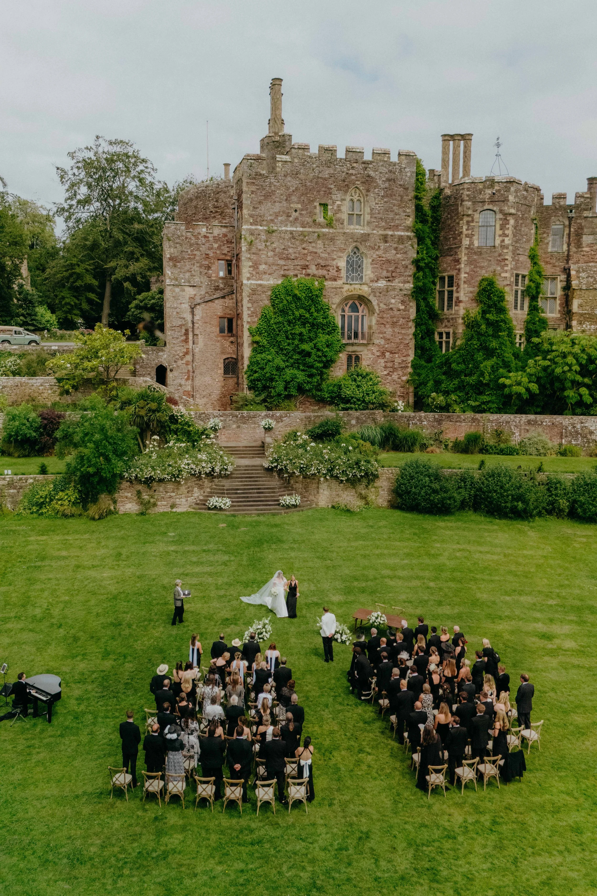 A wedding ceremony outdoors at Berkeley castle with a large gathering of guests seated on the grass, a bride and groom standing at the altar, and a band playing piano nearby.