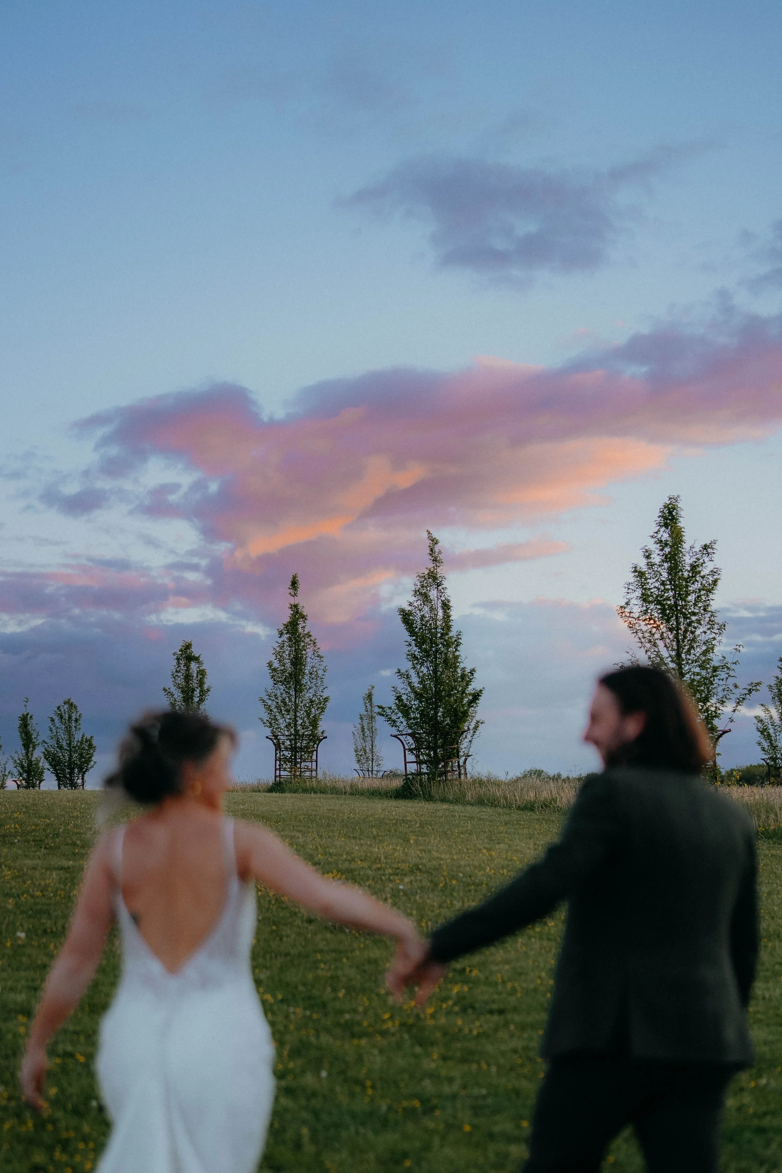 A couple holding hands outdoors in a grassy field at sunset, with a pastel-colored sky and trees in the background at Cripps Barn in the cotswolds