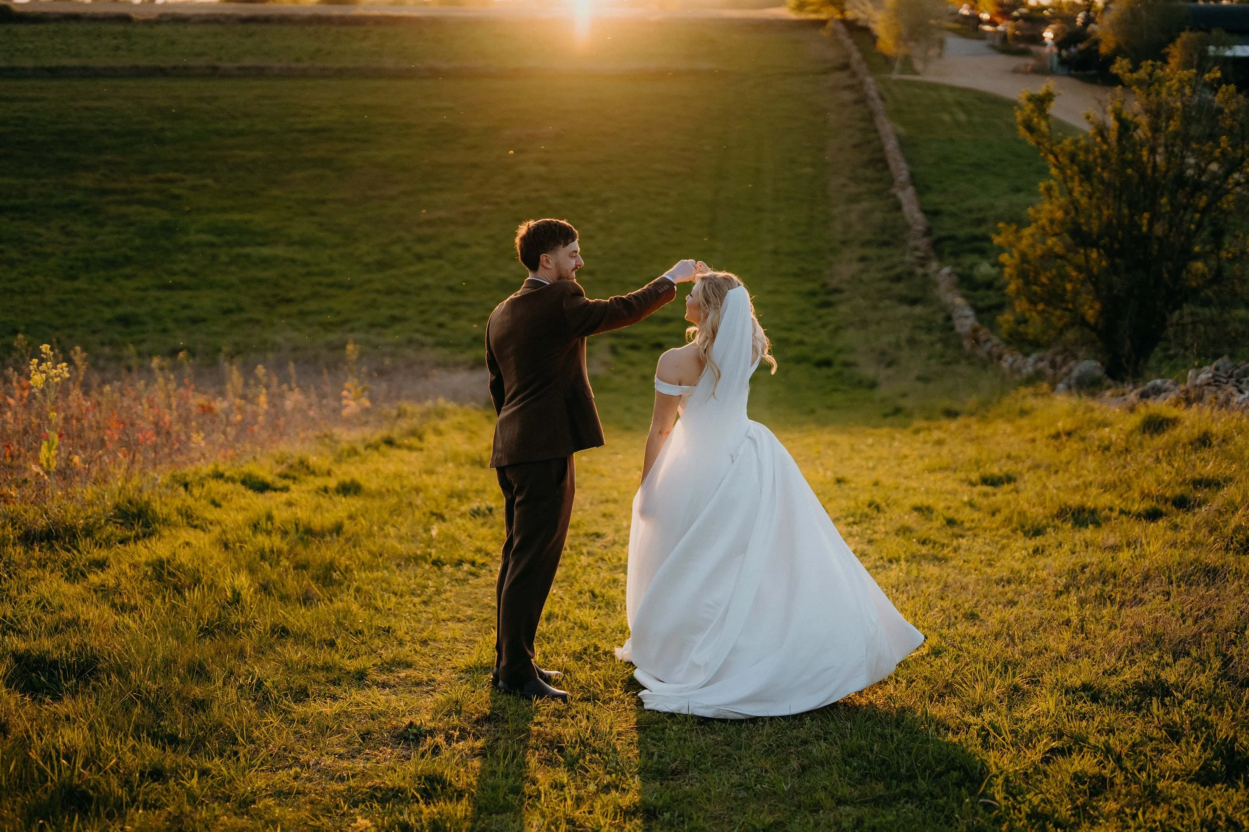 A couple dressed in wedding attire stands in a grassy area during sunset. The groom is pinning a flower on the bride's hair. The bride wears a white off-the-shoulder gown and a veil, while the groom wears a brown suit. The background features trees, 
