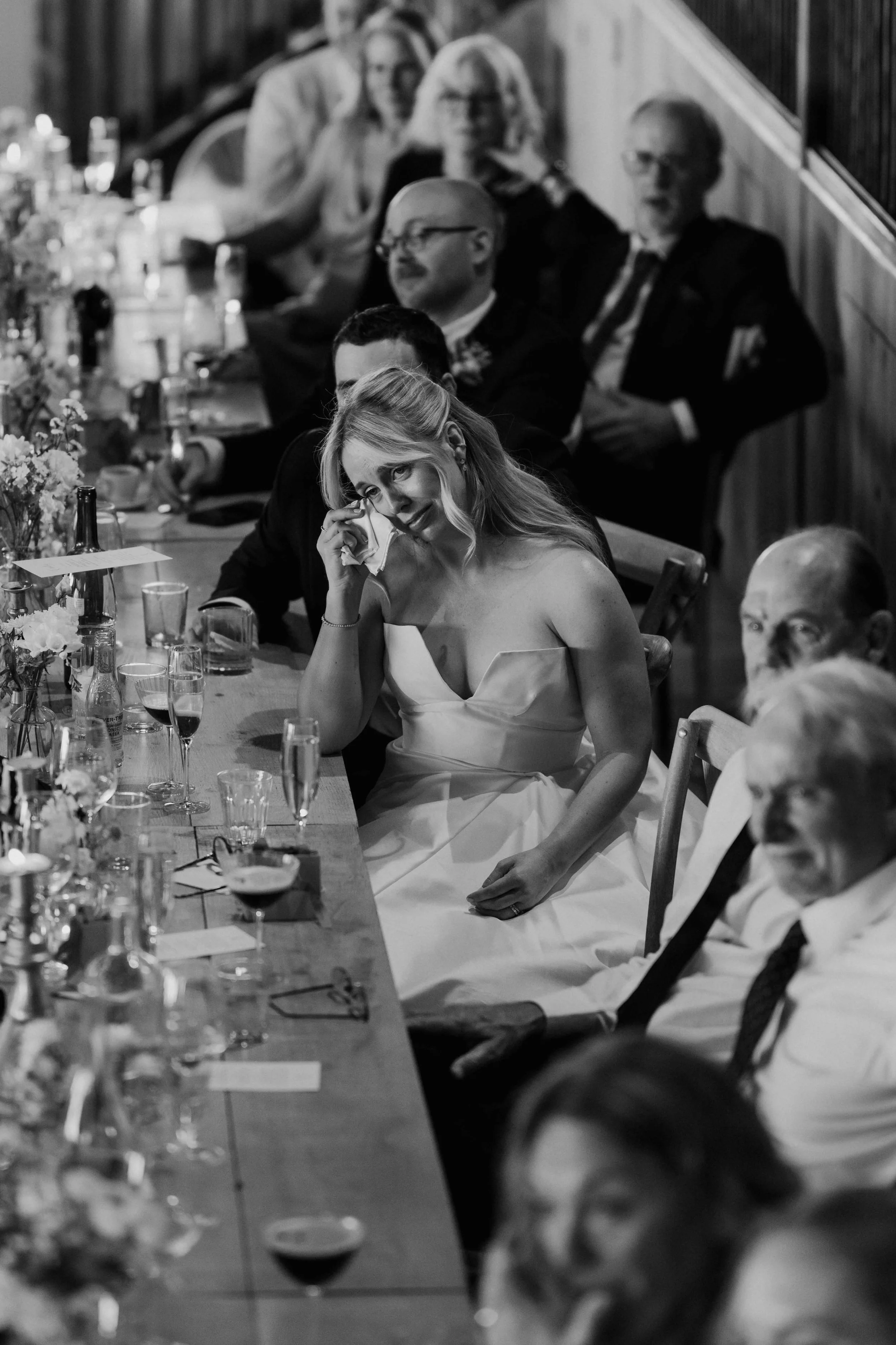 A black and white photo of a woman in a strapless dress wiping tears from her eyes during a formal event, sitting at a table with drinks and floral decorations, with other guests seated behind her.