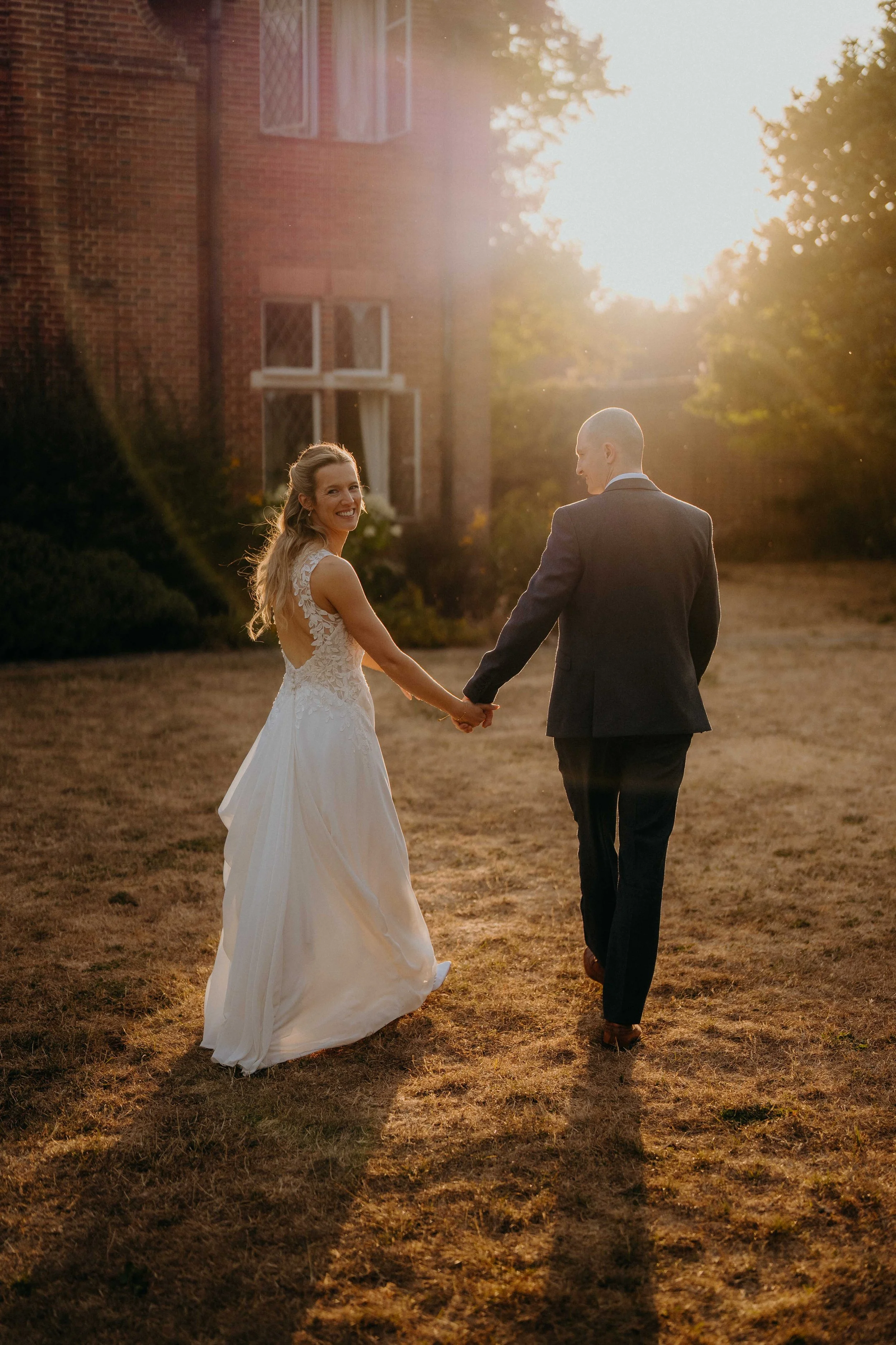 A couple, a bride in a white wedding dress and a groom in a dark suit, walking hand in hand outdoors at sunset, smiling and looking at each other.