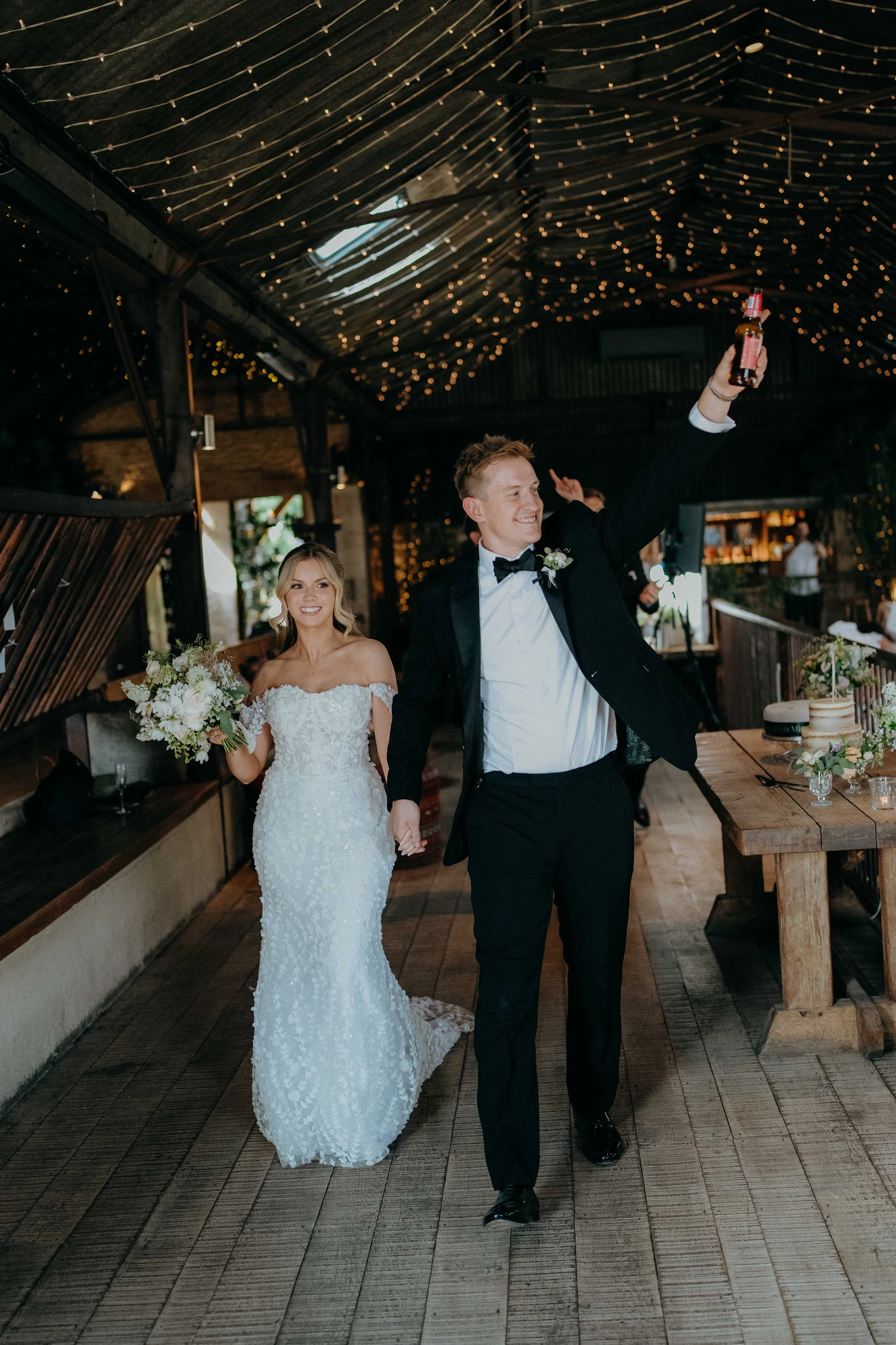 A bride and groom celebrating at their wedding reception indoors with string lights on the ceiling. The bride is holding a bouquet of white flowers and wearing a white lace wedding gown. The groom is wearing a black tuxedo and is raising a bottle in 