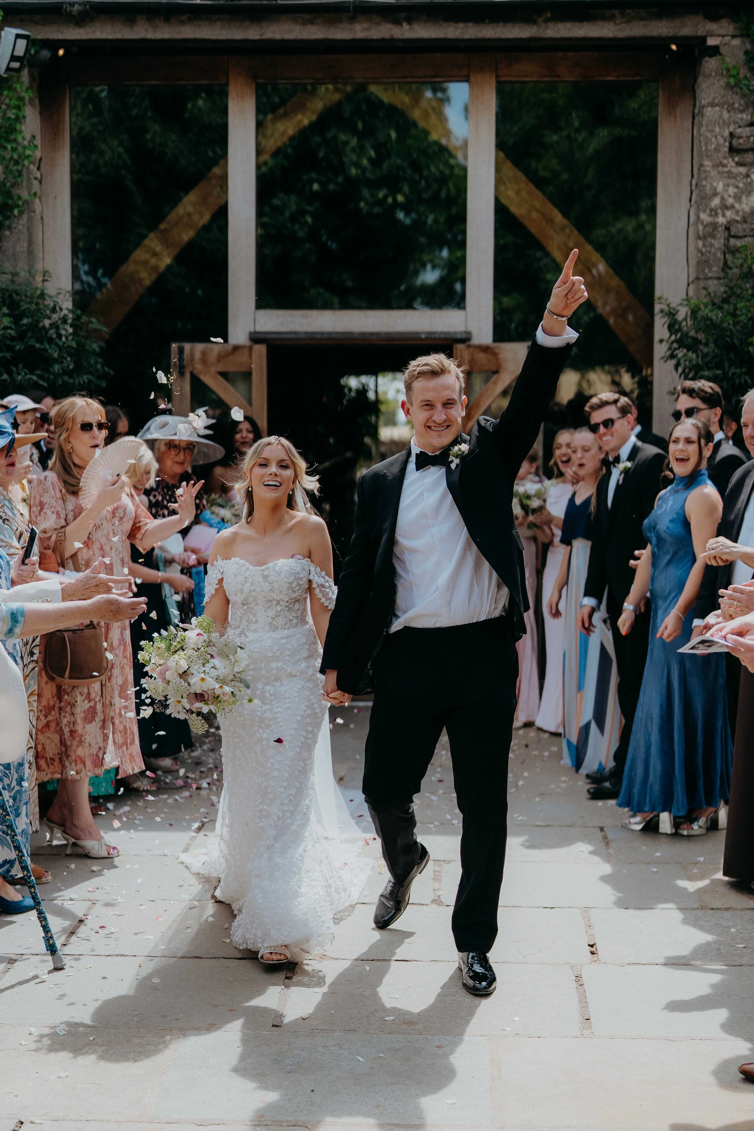 A newlywed couple walks hand in hand outdoors as guests cheer and throw flower petals at Stone Barn. The groom, dressed in a black tuxedo, points upward and smiles, while the bride, in a white lace wedding gown, holds a bouquet and also smiles. 