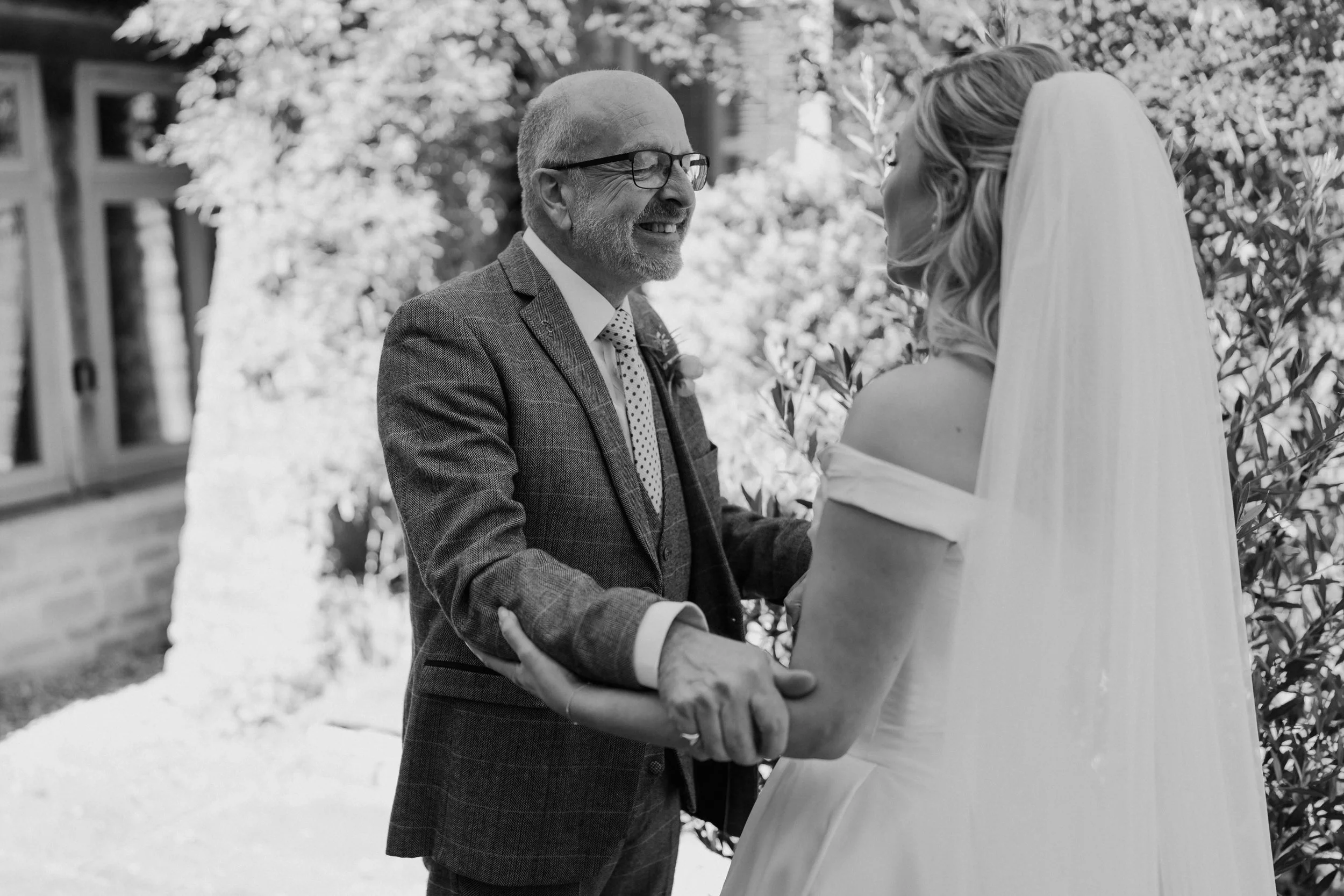 An older man with glasses and a beard, wearing a suit, smiling, during a wedding ceremony. A bride with long hair and a veil is holding his hands, standing outdoors surrounded by trees.