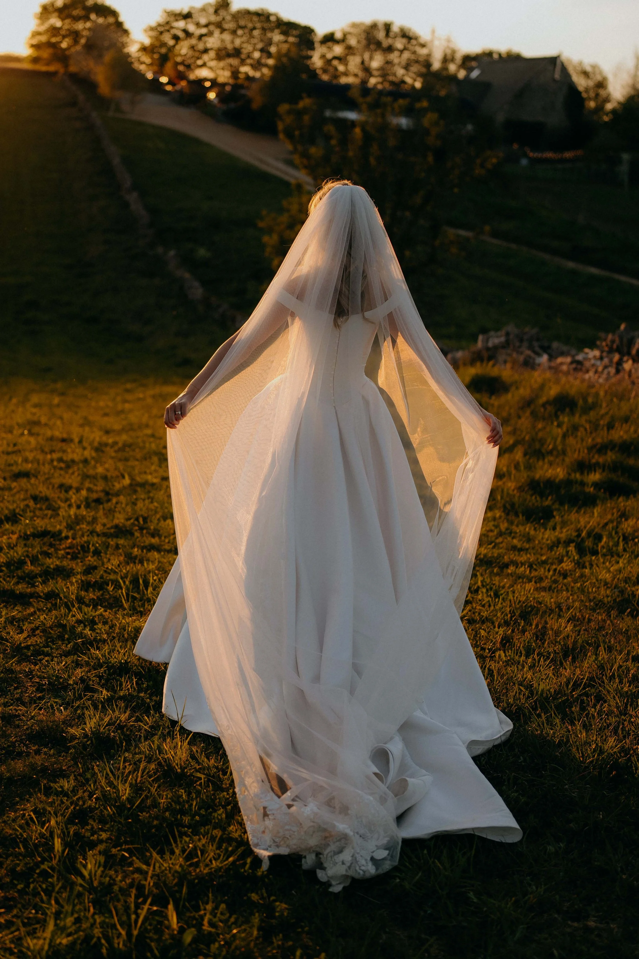 A woman in an elegant white wedding dress and veil standing outdoors at sunset on a grassy field at Stone Barn in the cotswolds with trees and houses in the background.