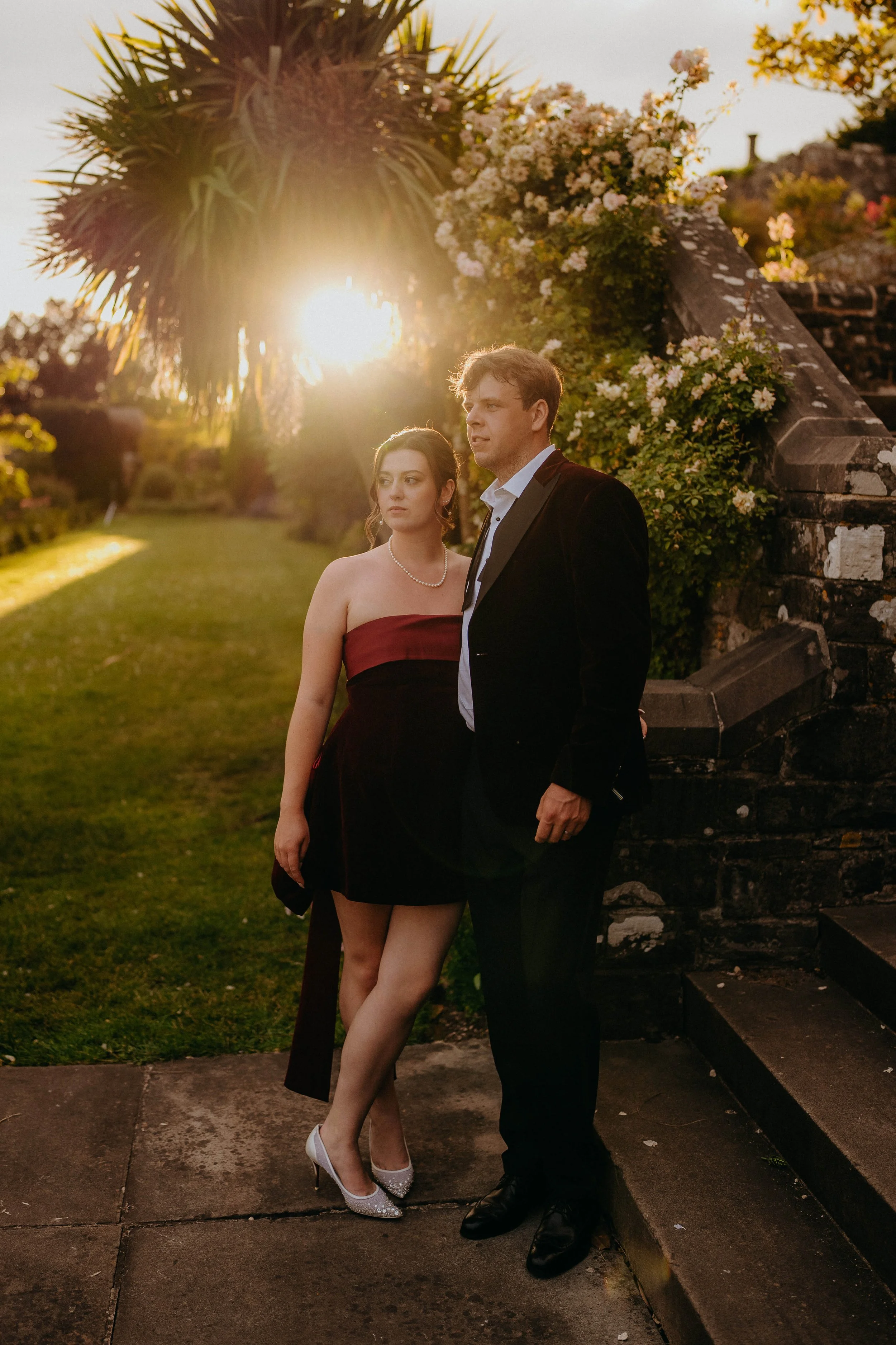 A young couple dressed formally standing outdoors on a sidewalk with a stone staircase covered in flowers behind them. The woman is wearing a strapless maroon dress with a pearl necklace and white high heels, and the man is in a tuxedo with a white s