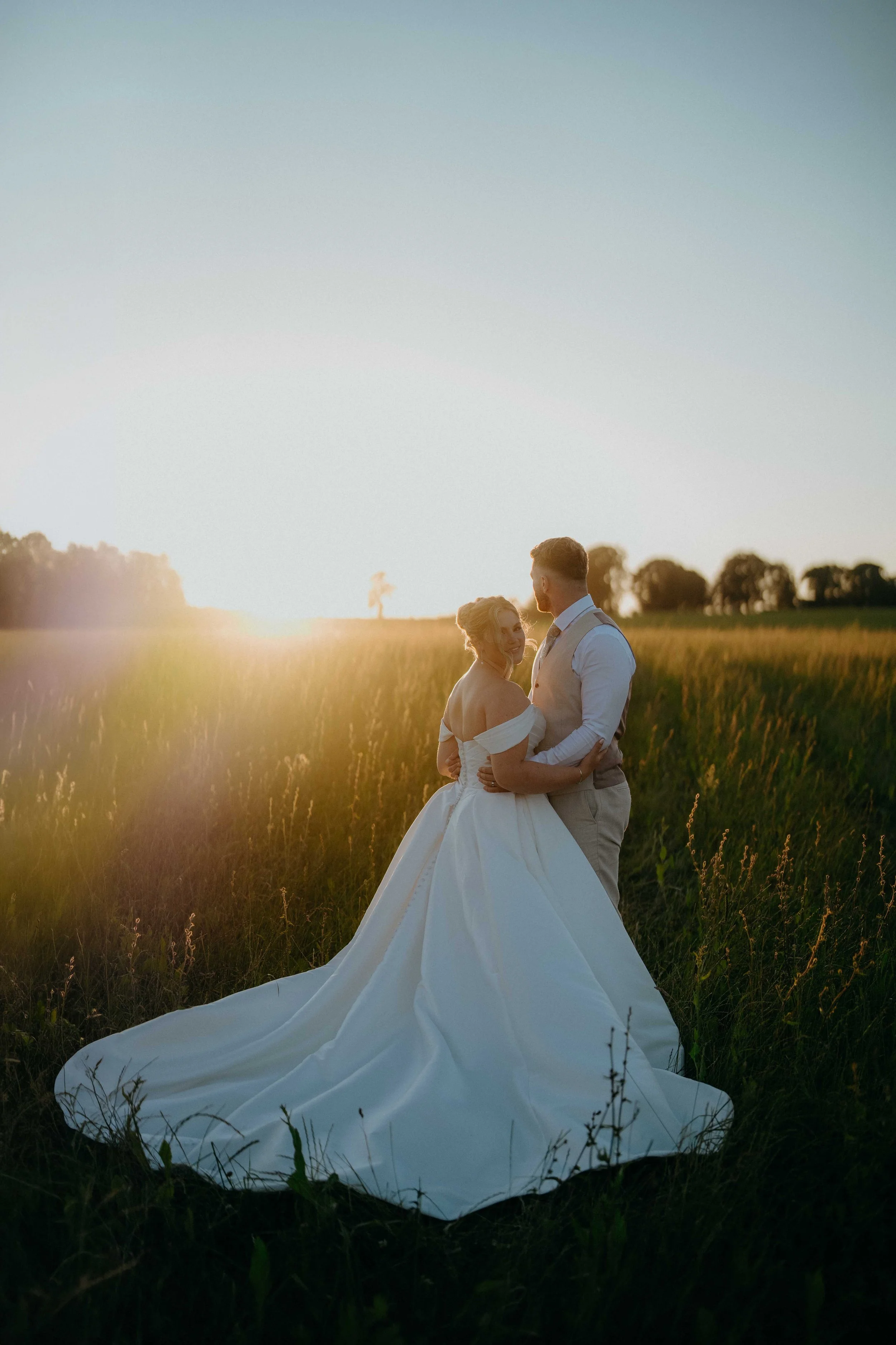 A bride and groom standing in a field during sunset, embracing each other, with the bride wearing a white wedding gown and the groom in a beige vest and shirt.