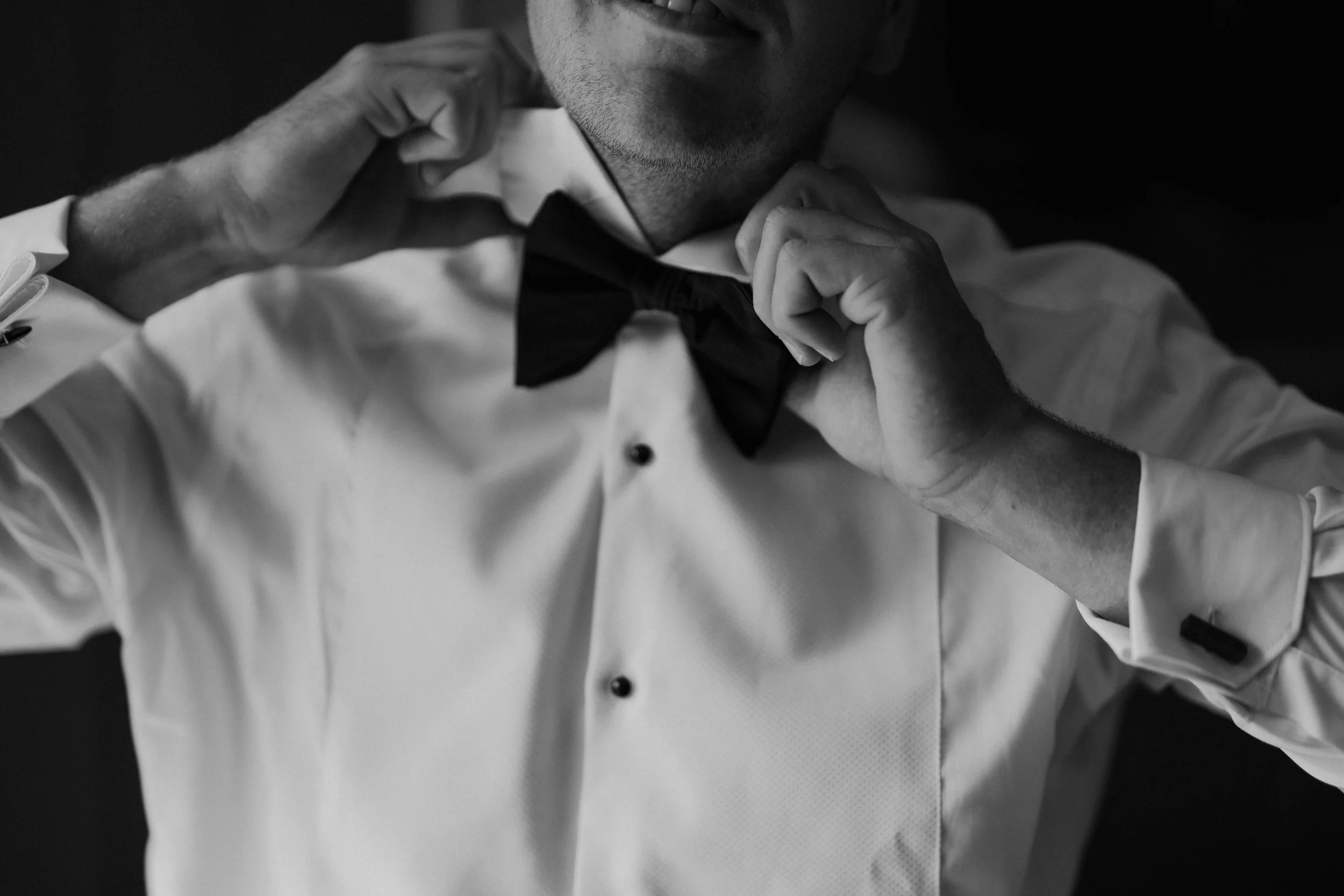 A man in a white dress shirt adjusts his black bow tie. The image is in black and white, capturing his face partially and emphasizing his hands preparing his attire.