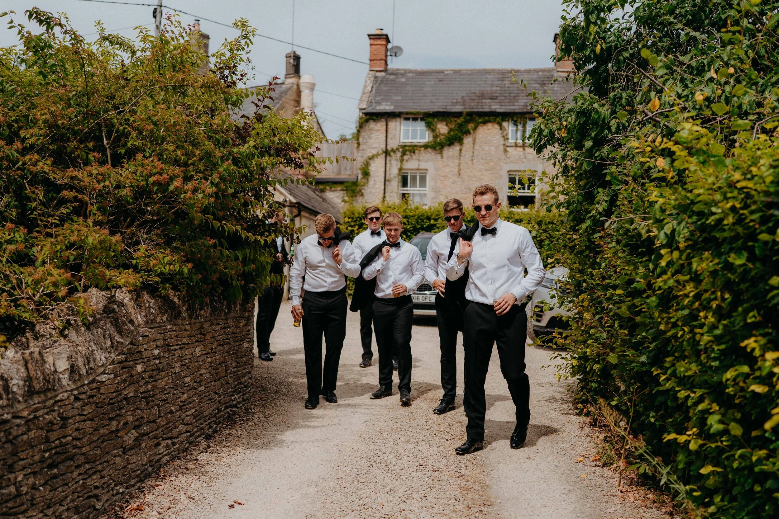 A group of six men in suits and sunglasses walking down a narrow alleyway with green bushes on either side, in front of houses with stone walls and chimneys.