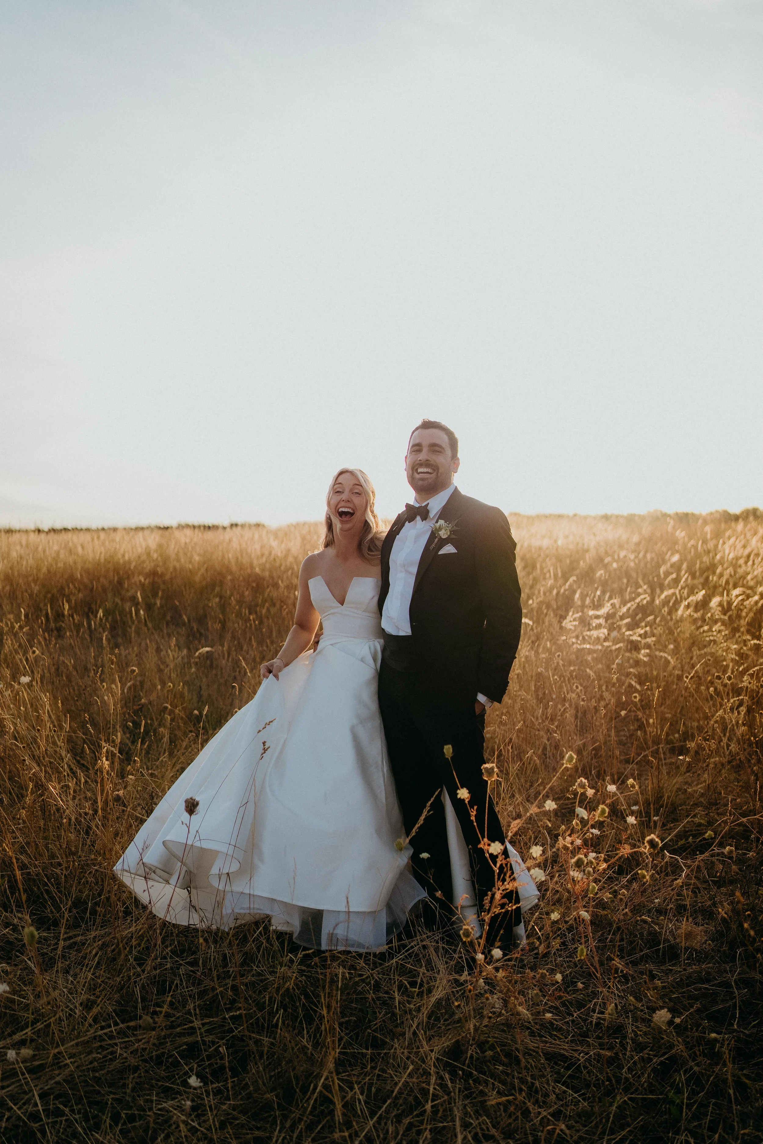 A smiling bride and groom standing in a field at sunset, the bride wearing a white wedding dress and the groom in a black tuxedo at Stone Barn in the cotswolds