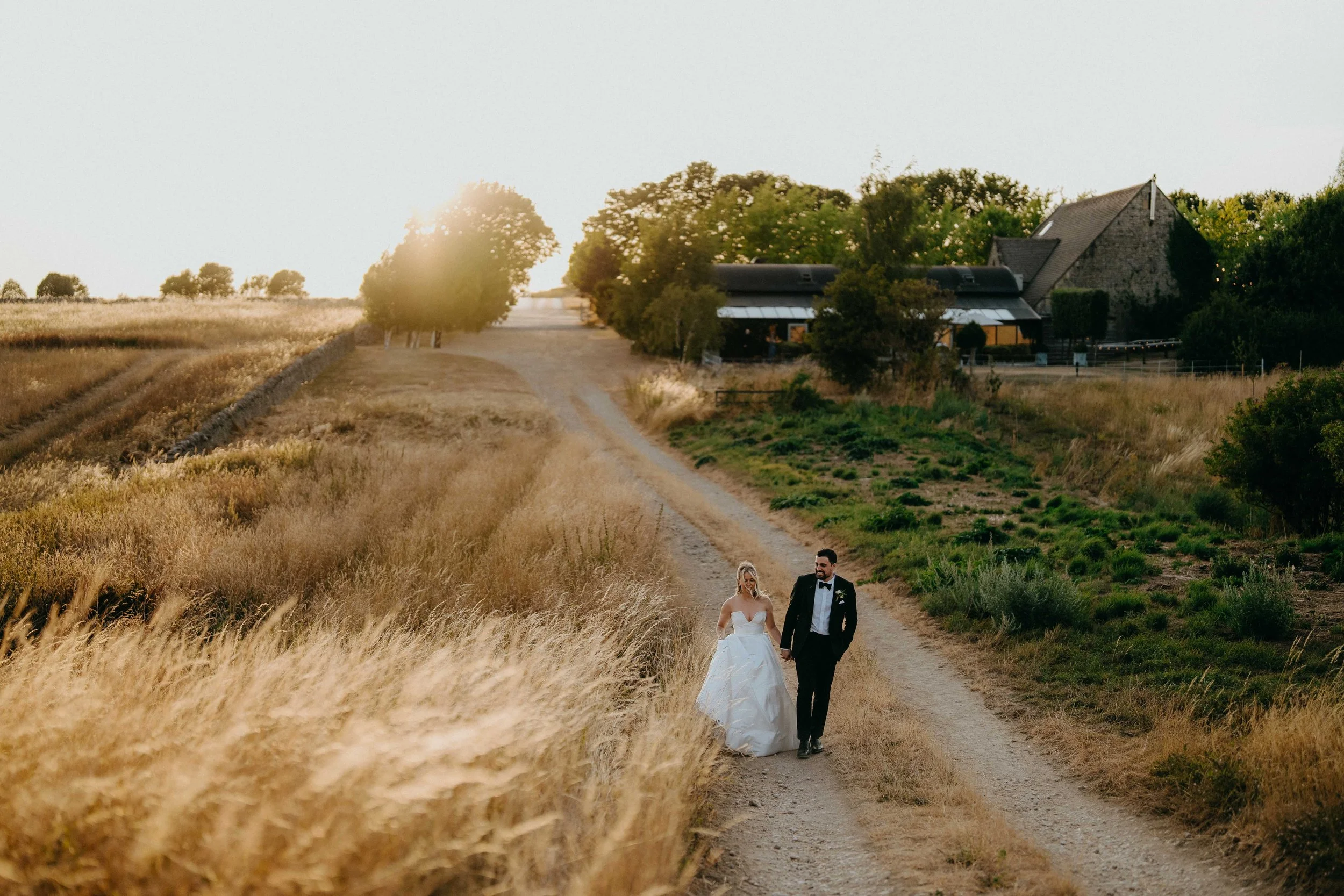 A bride and groom walking hand in hand along a dirt path in a rural field at sunset, with trees at Stone Barn in the cotswolds in the background.