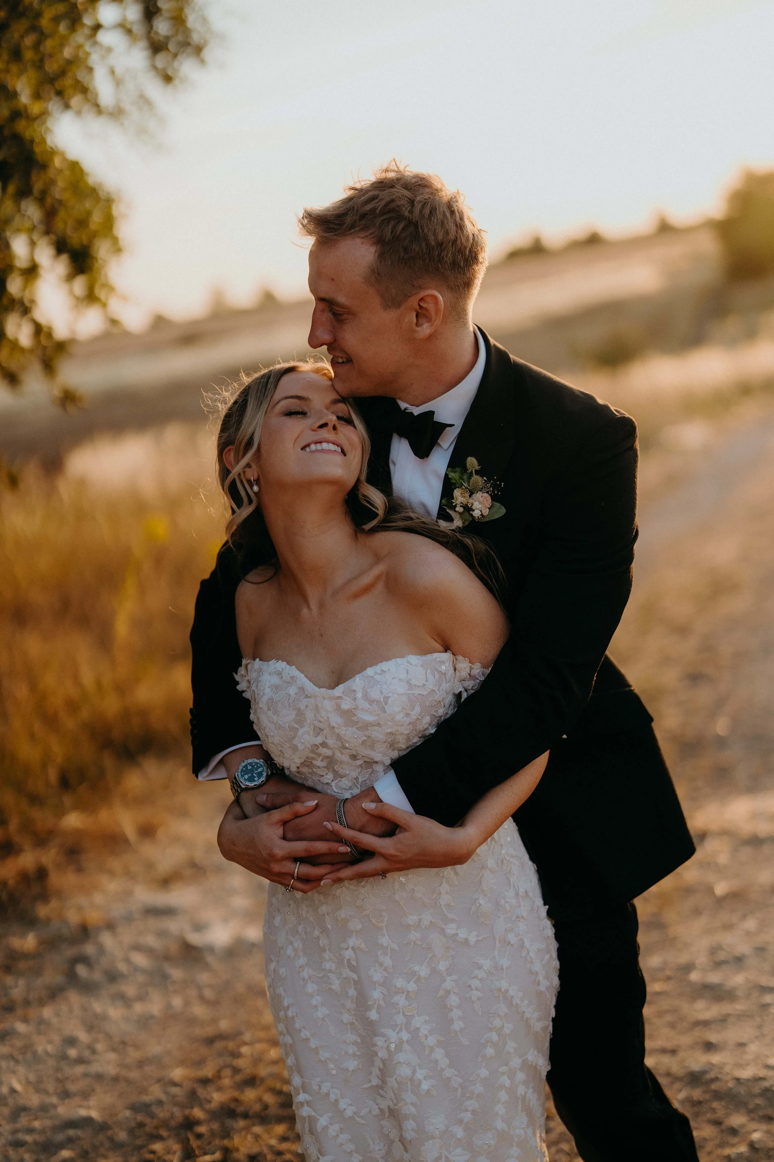 A couple dressed in wedding attire, smiling and embracing outdoors during sunset, with a blurred landscape and trees in the background at Stone Barn in the cotswolds
