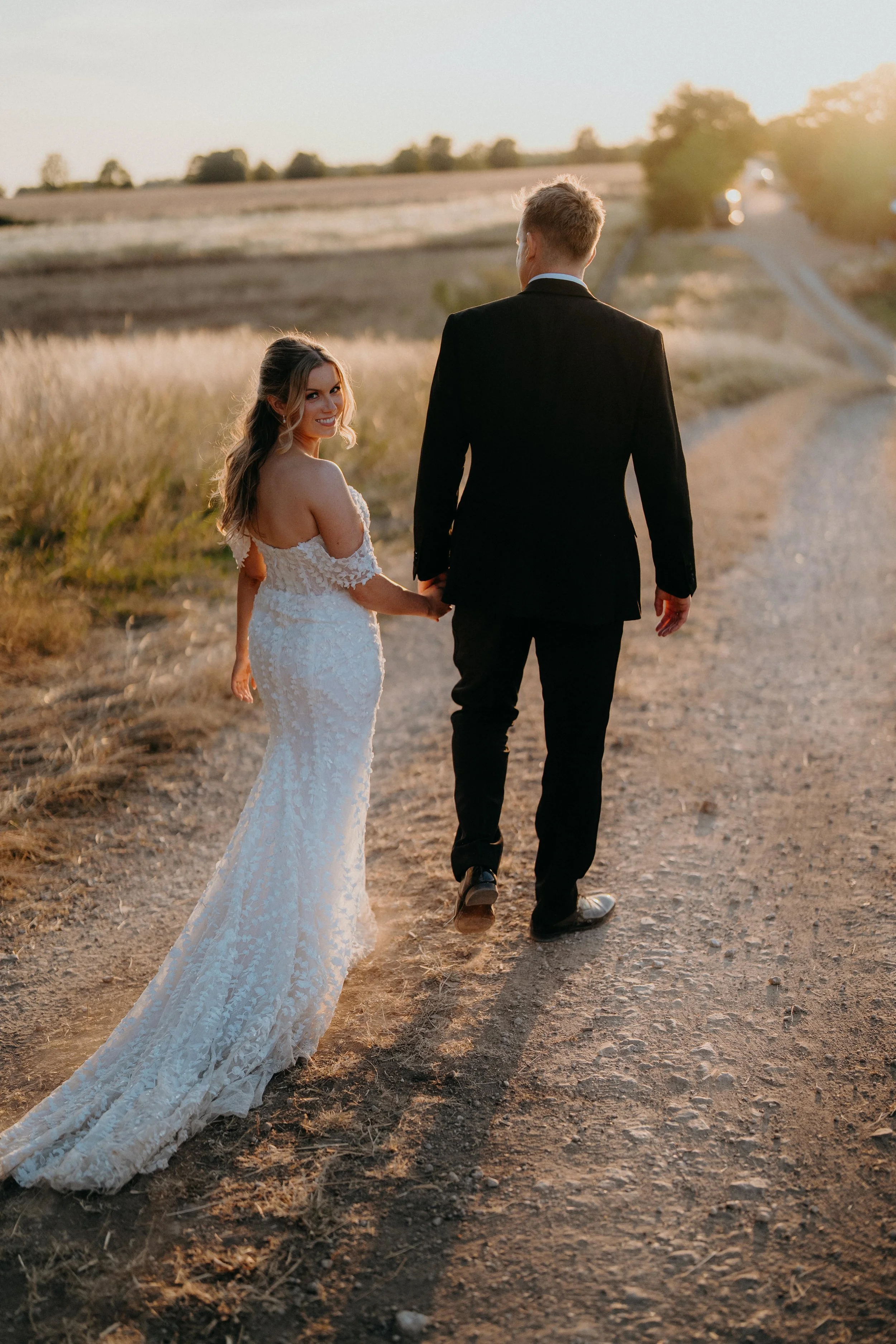 A happy bride in a white lace wedding dress holding hands with a groom in a black suit walking on a dirt path in a rural area during sunset.