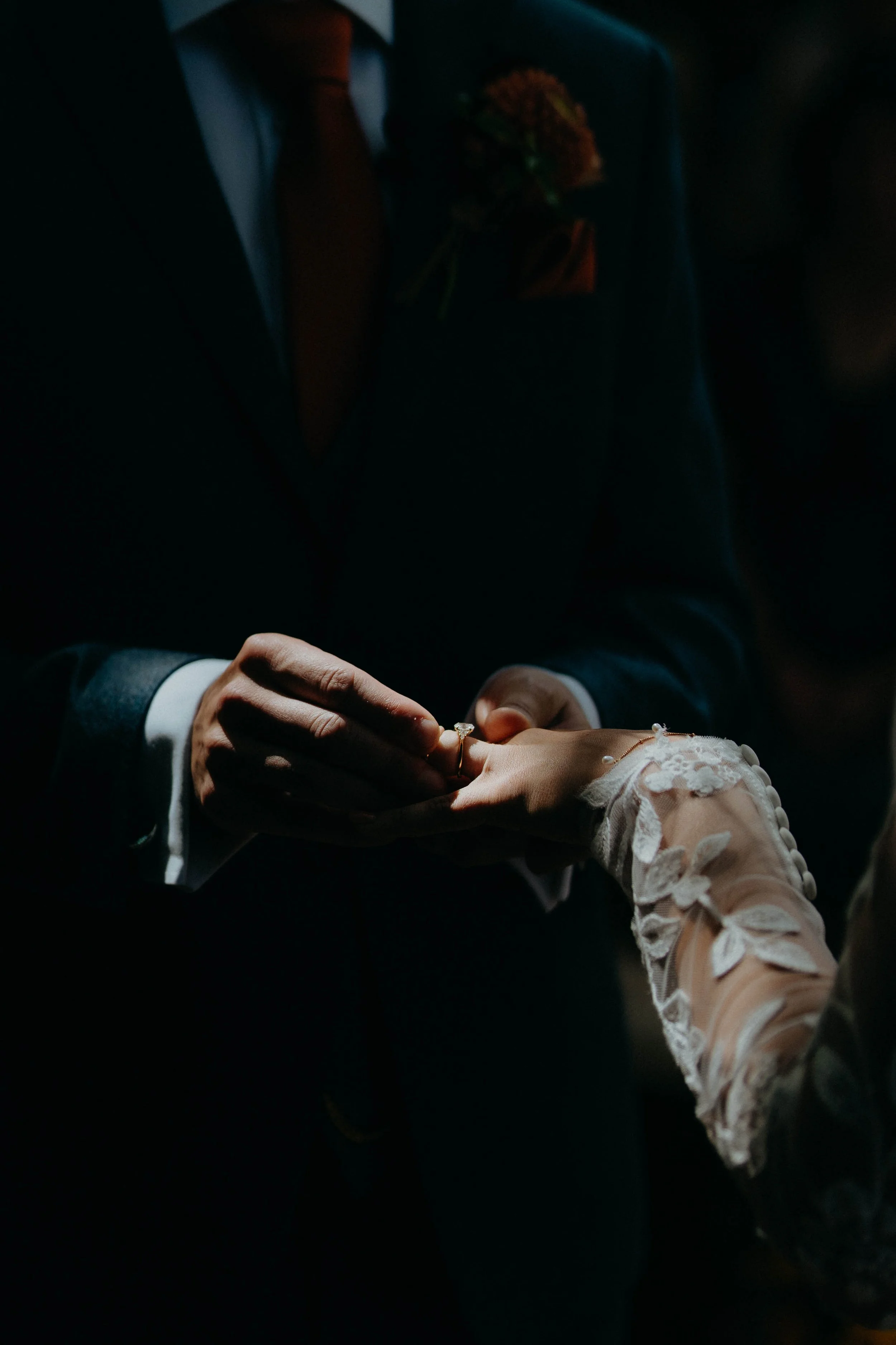 A couple exchanging wedding rings during a wedding ceremony, with the groom dressed in a dark suit and the bride wearing a dress with lace sleeve details.