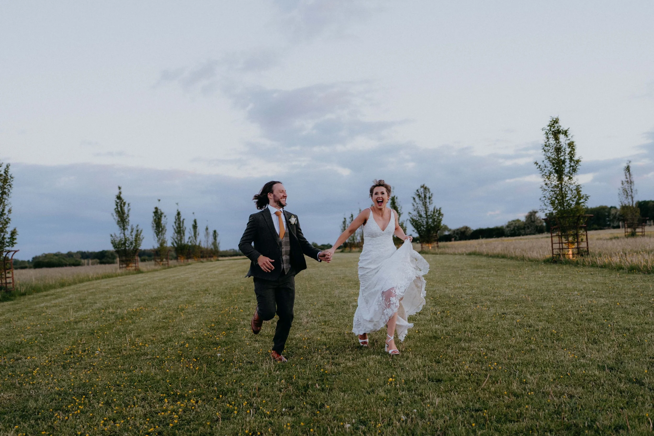 A bride and groom running hand in hand on a grassy field outdoors during cloudy weather, smiling and joyful at Cripps Barn in the cotswolds
