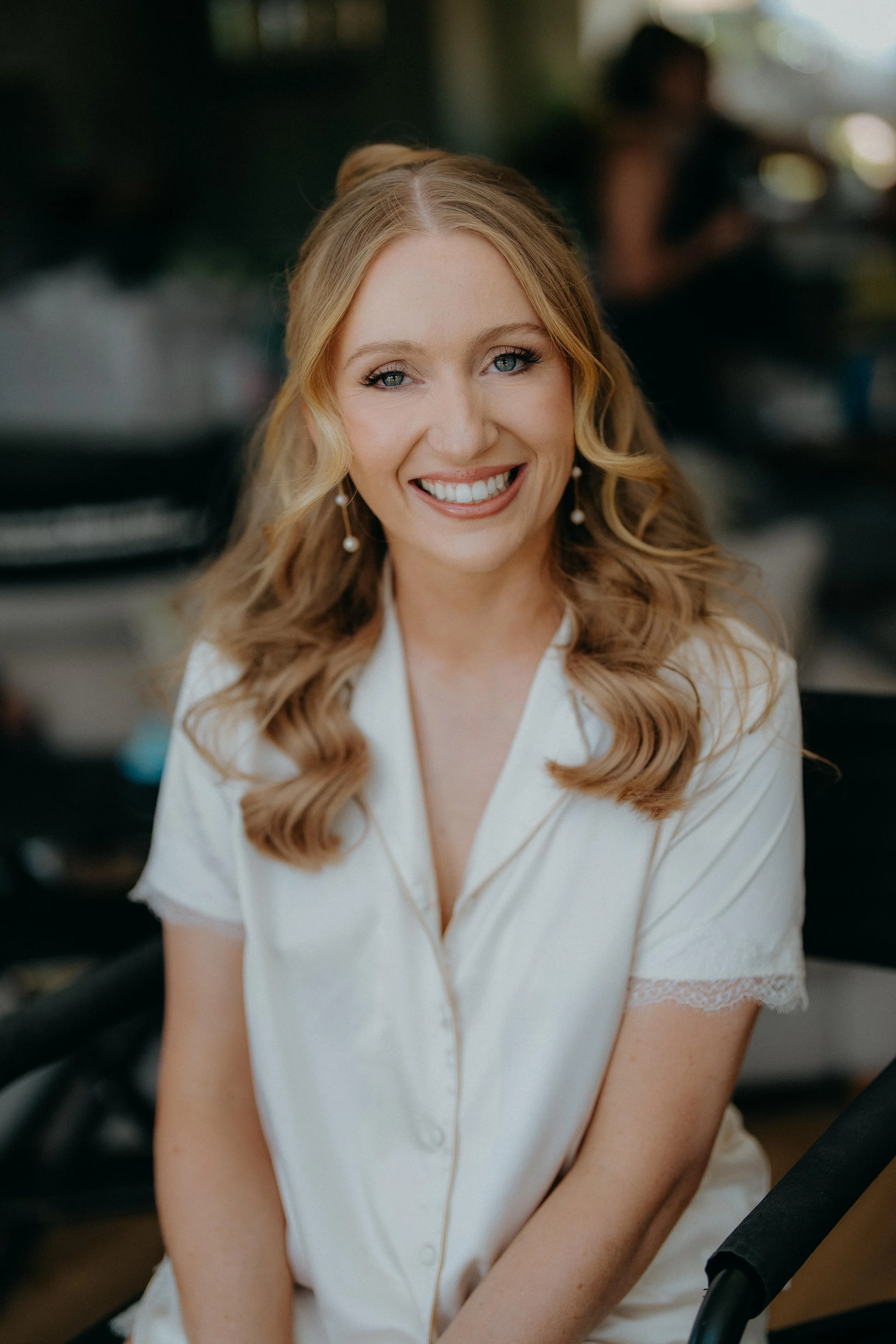 A smiling woman with blonde hair styled in loose waves, wearing pearl earrings and a cream-colored top with lace trim on the sleeves, sitting in an indoor setting with blurred background.