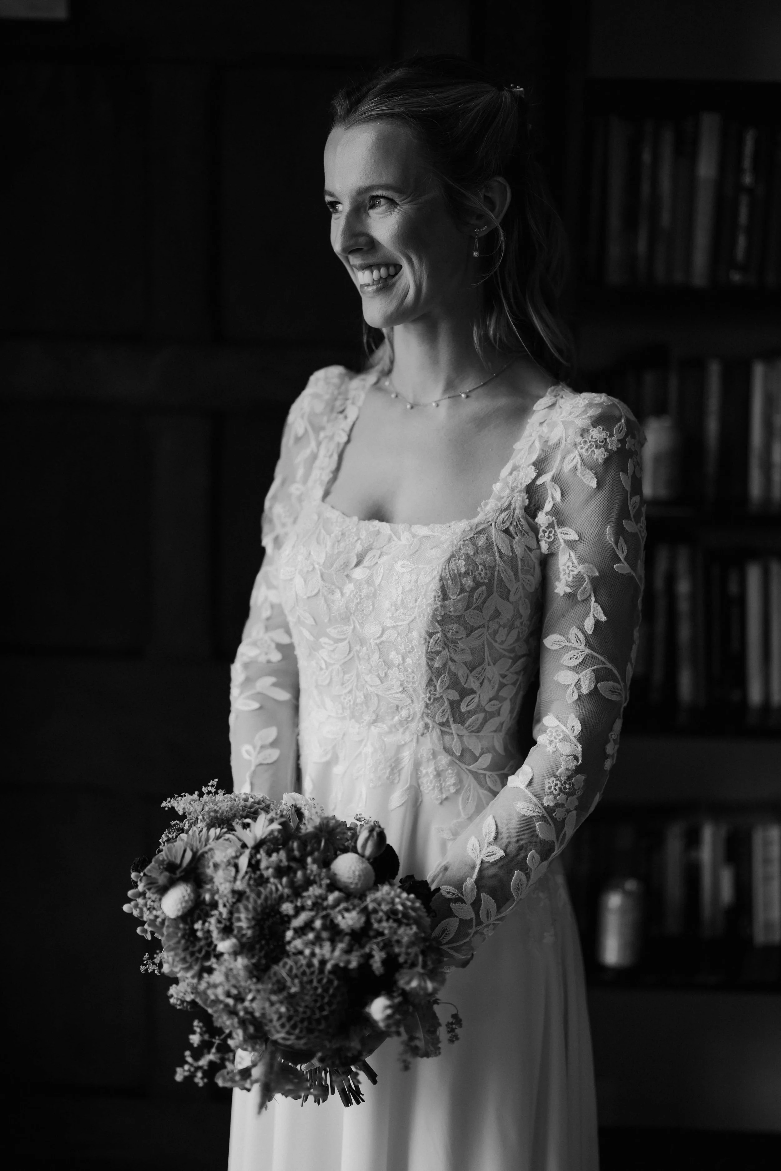 A smiling bride in a lace wedding dress holding a bouquet of flowers in a room with dark wood bookshelves.