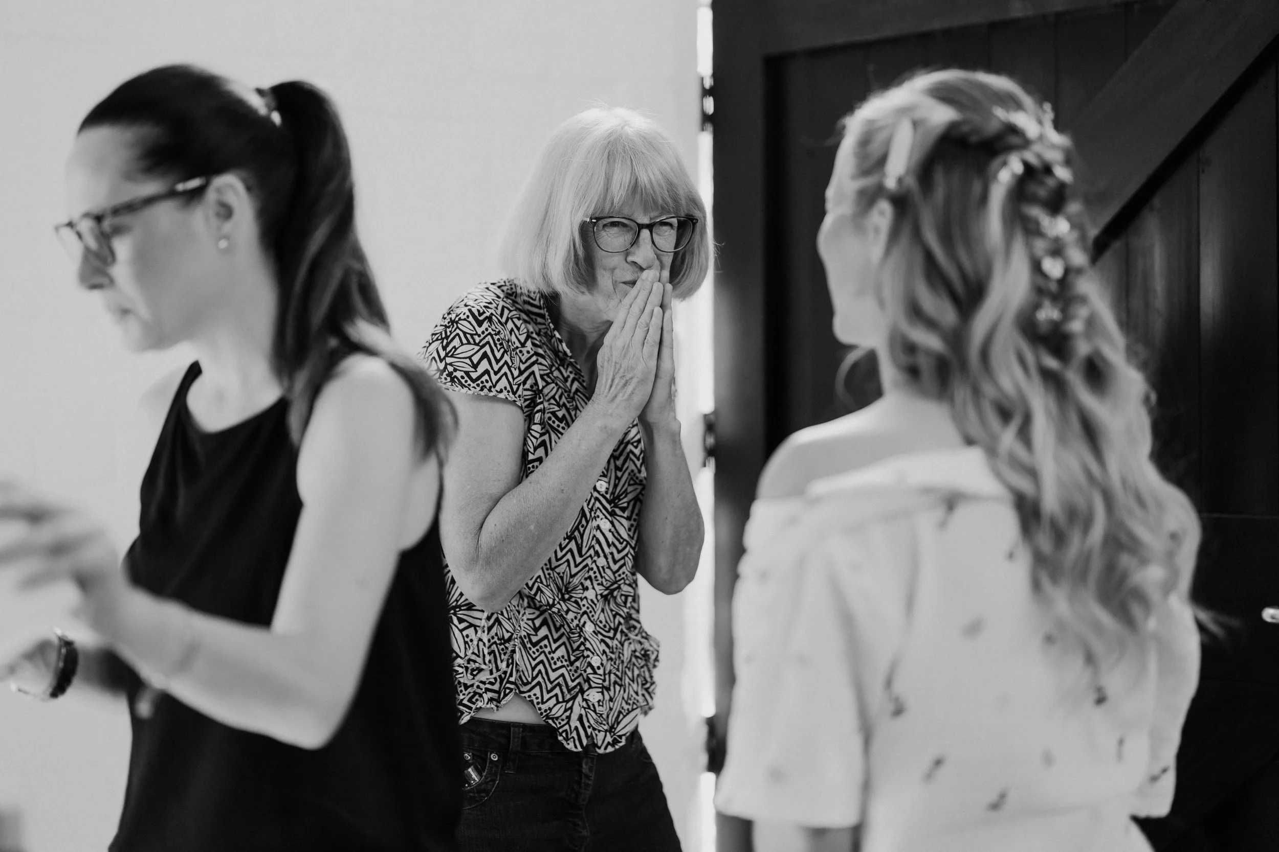 An older woman with glasses and light-colored hair, wearing a patterned top, appears to be praying with her hands clasped near her face. She is surrounded by young women, some with braided hairstyles, wearing formal or semi-formal attire. The scene s