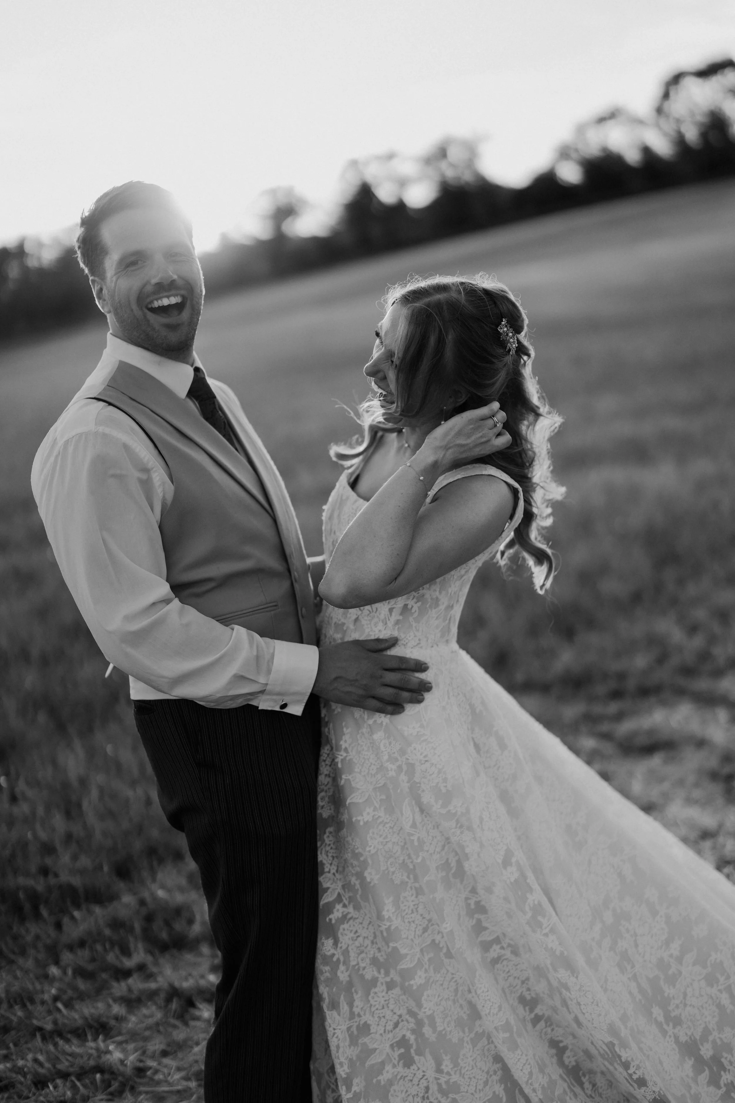 A black-and-white photo of a happy couple dressed in wedding attire, standing in a field with trees in the background. The man is laughing, wearing a vest and tie, and the woman is smiling, in a lace wedding dress, touching her hair.