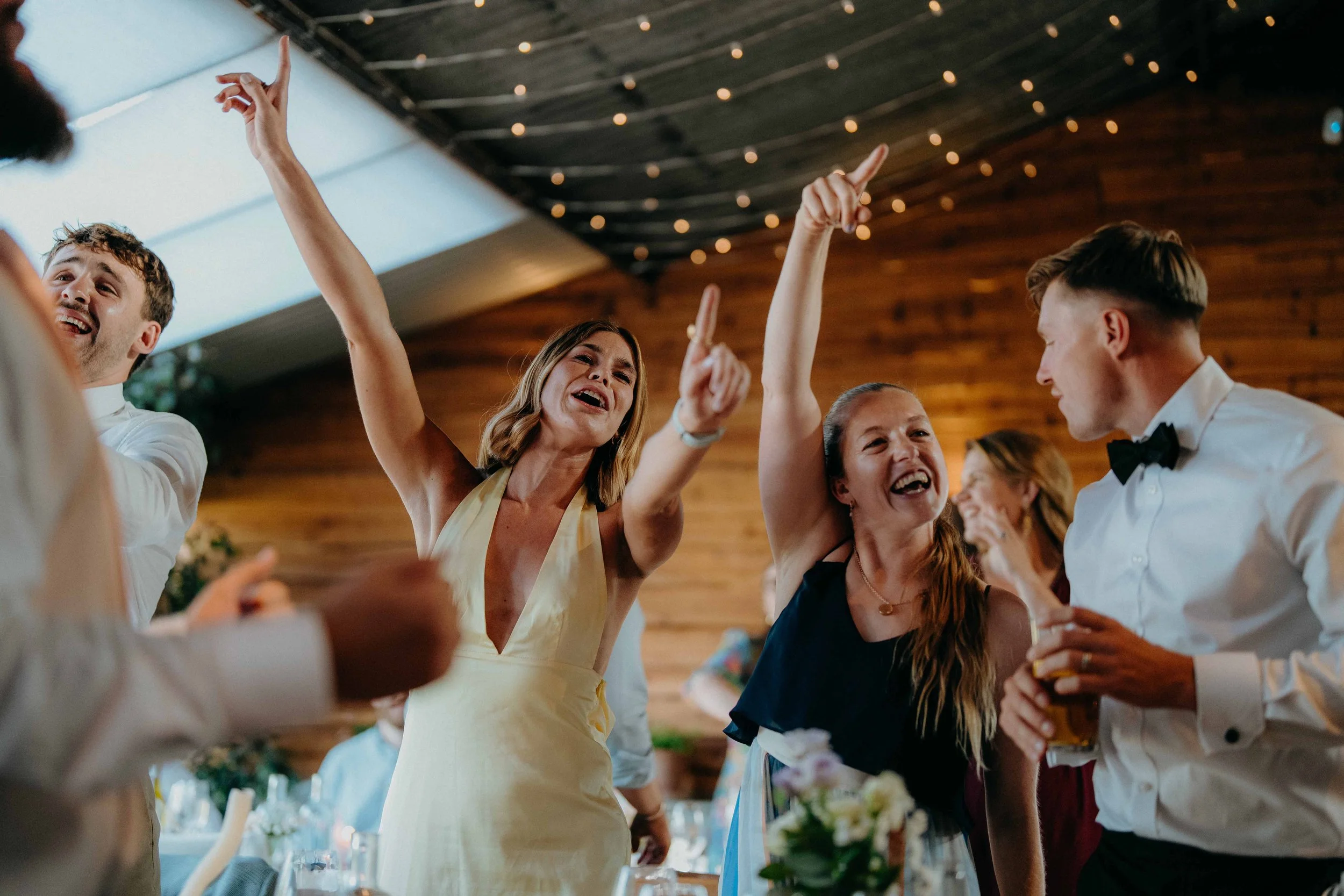 People dancing and celebrating at a wedding reception in a venue with string lights and wooden walls at a singing waiter performance at Stone Barn in the cotswolds