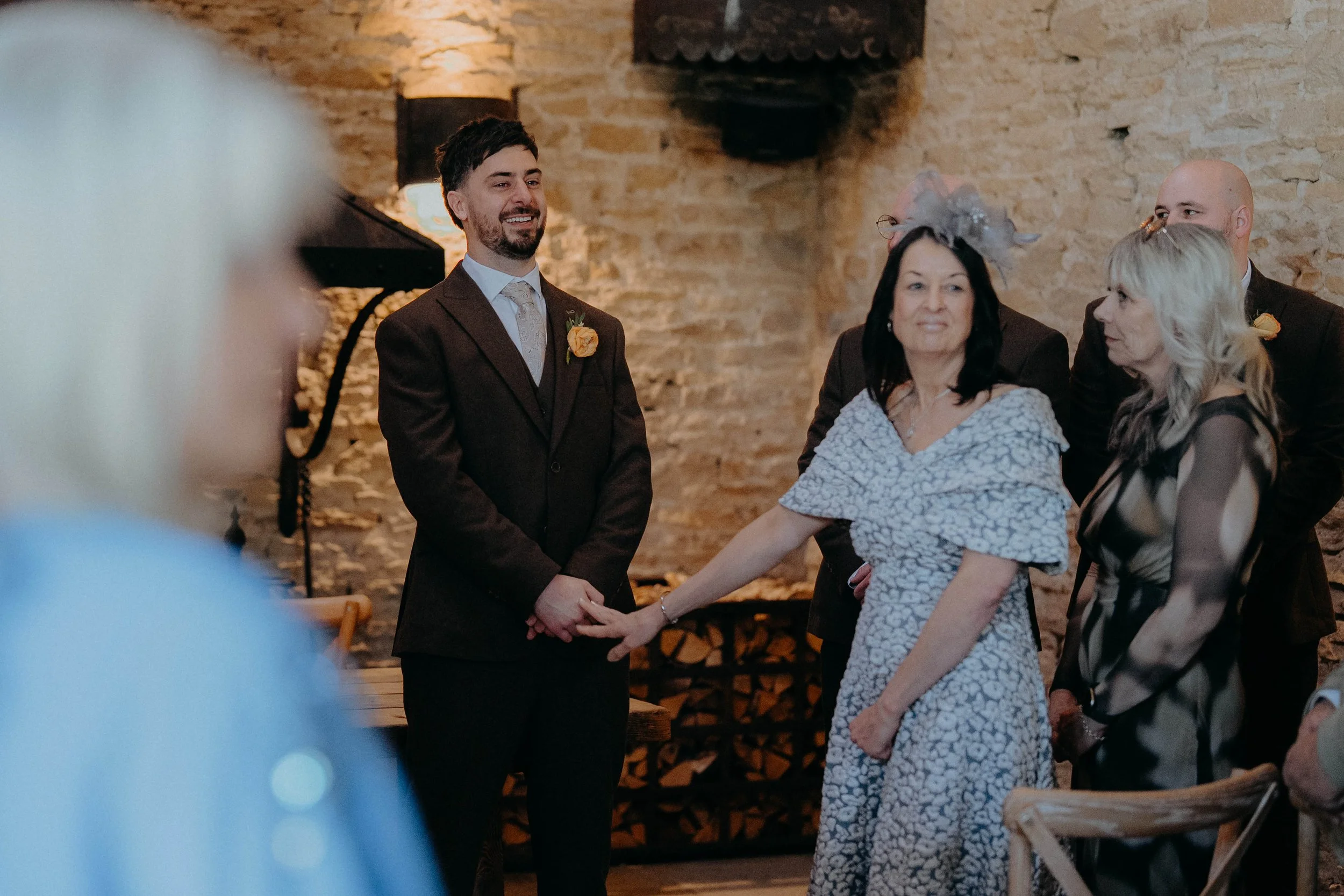 A groom and bride holding hands and exchanging vows during a wedding ceremony in a rustic indoor setting with exposed brick walls. The groom has dark hair and a beard, wearing a dark brown suit with a yellow boutonnière. The bride has blonde hair, we