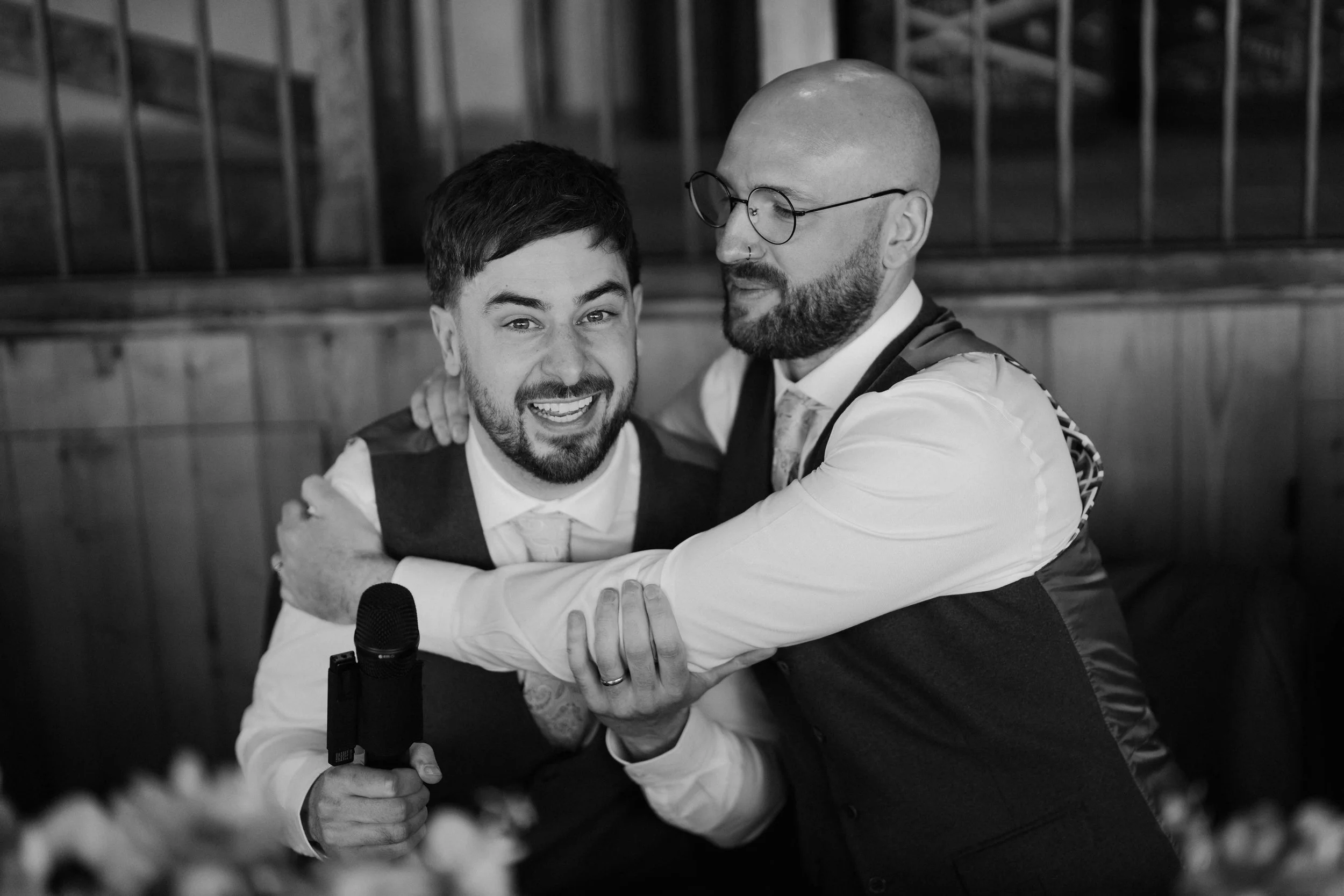 Two men embracing each other in a hug, smiling, on a stage with a microphone in front of them, with a blurred background of flowers or plants.