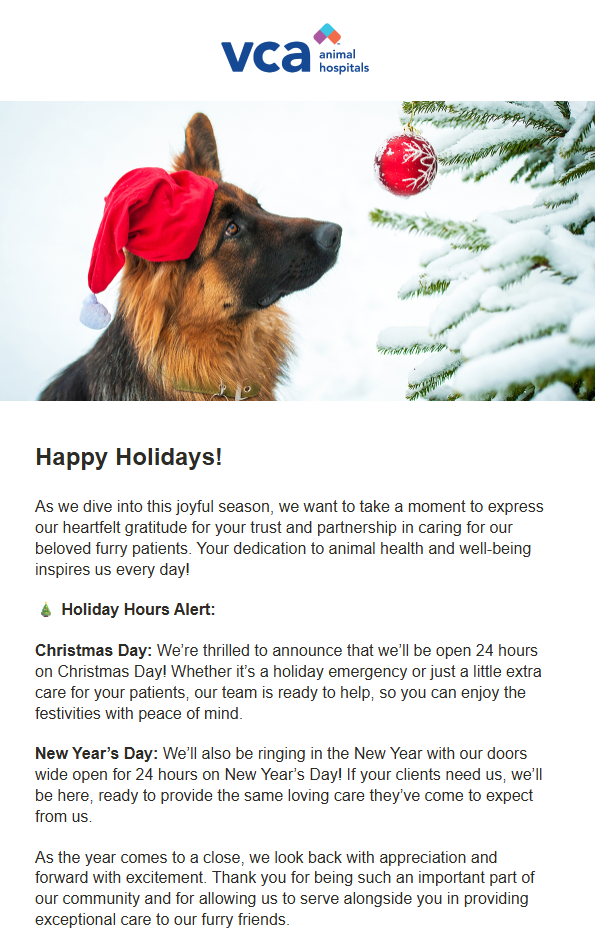 A German shepherd dog wearing a red Santa hat with a white pom-pom looking at a red Christmas ornament hanging on a snow-covered Christmas tree, with a snowy background.