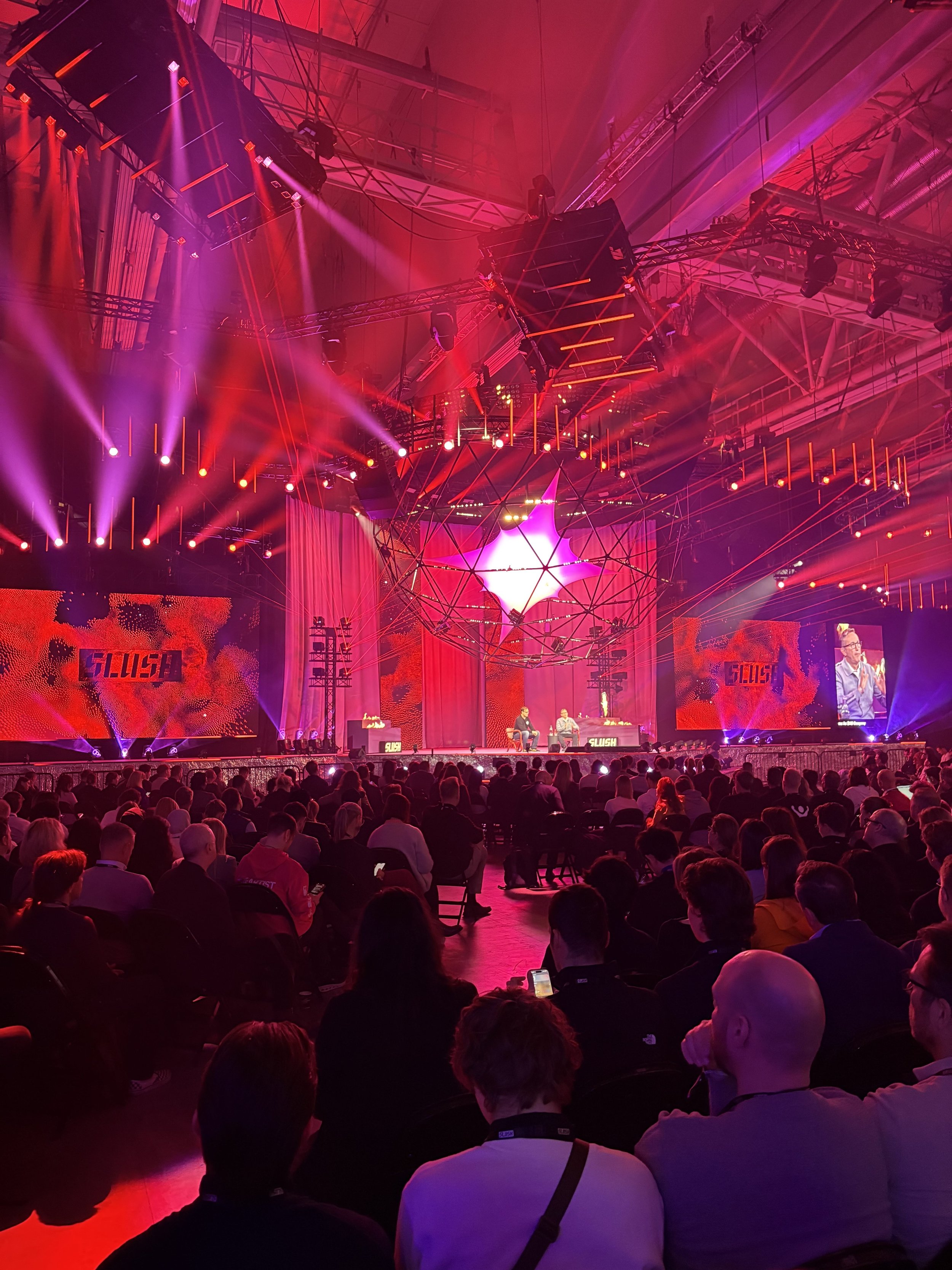 A large indoor conference or event with a seated audience facing a stage with speakers, monitors, and colorful lighting, including a star-shaped light fixture hanging from the ceiling and large screens displaying a speaker and graphics.