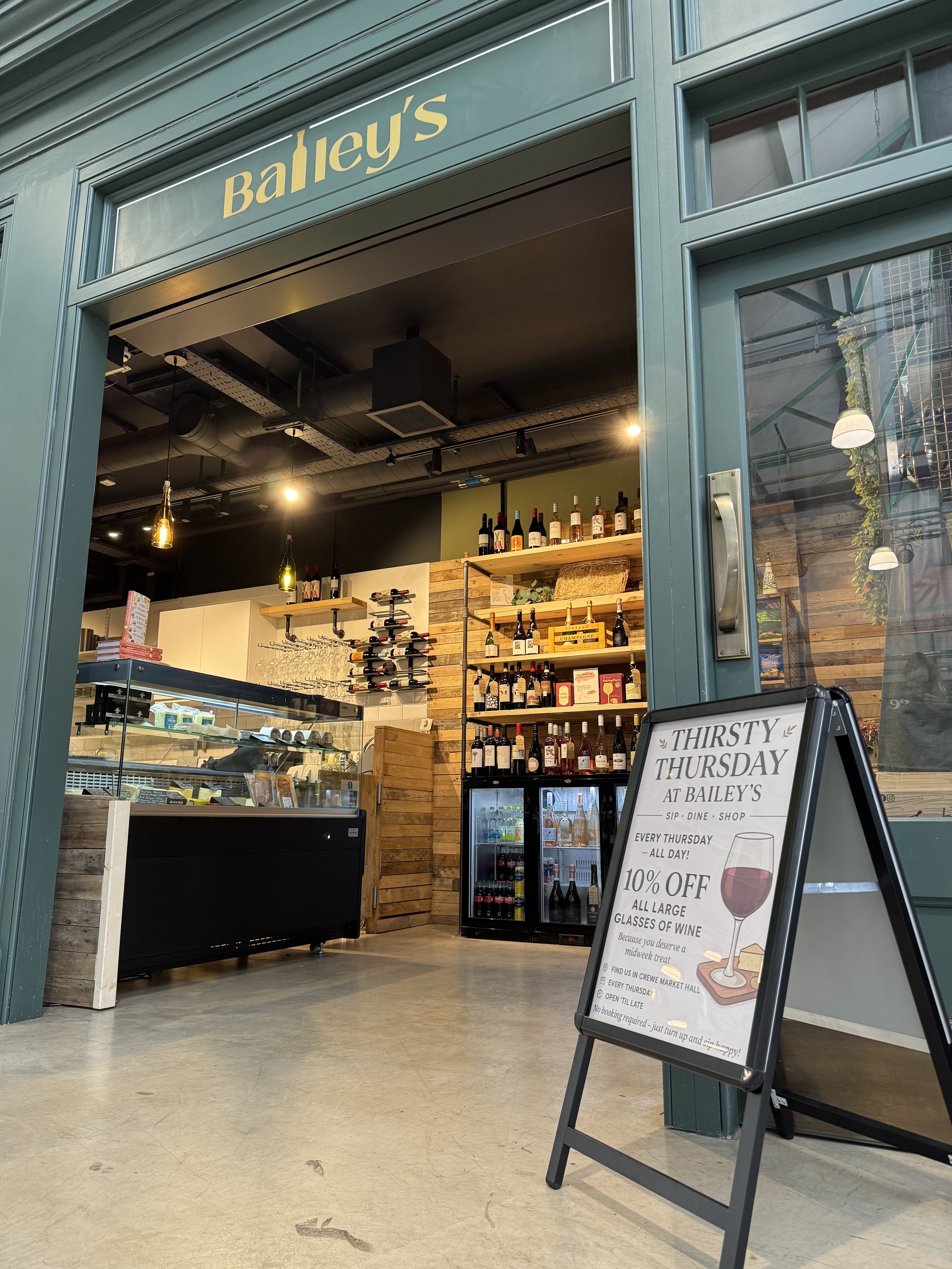 A wine shop named Bailey's with a wooden interior, displaying bottles on shelves and a glass counter with wine-related items. An A-frame sign outside advertises "Thirsty Thursday" with 10% off large glasses of wine.
