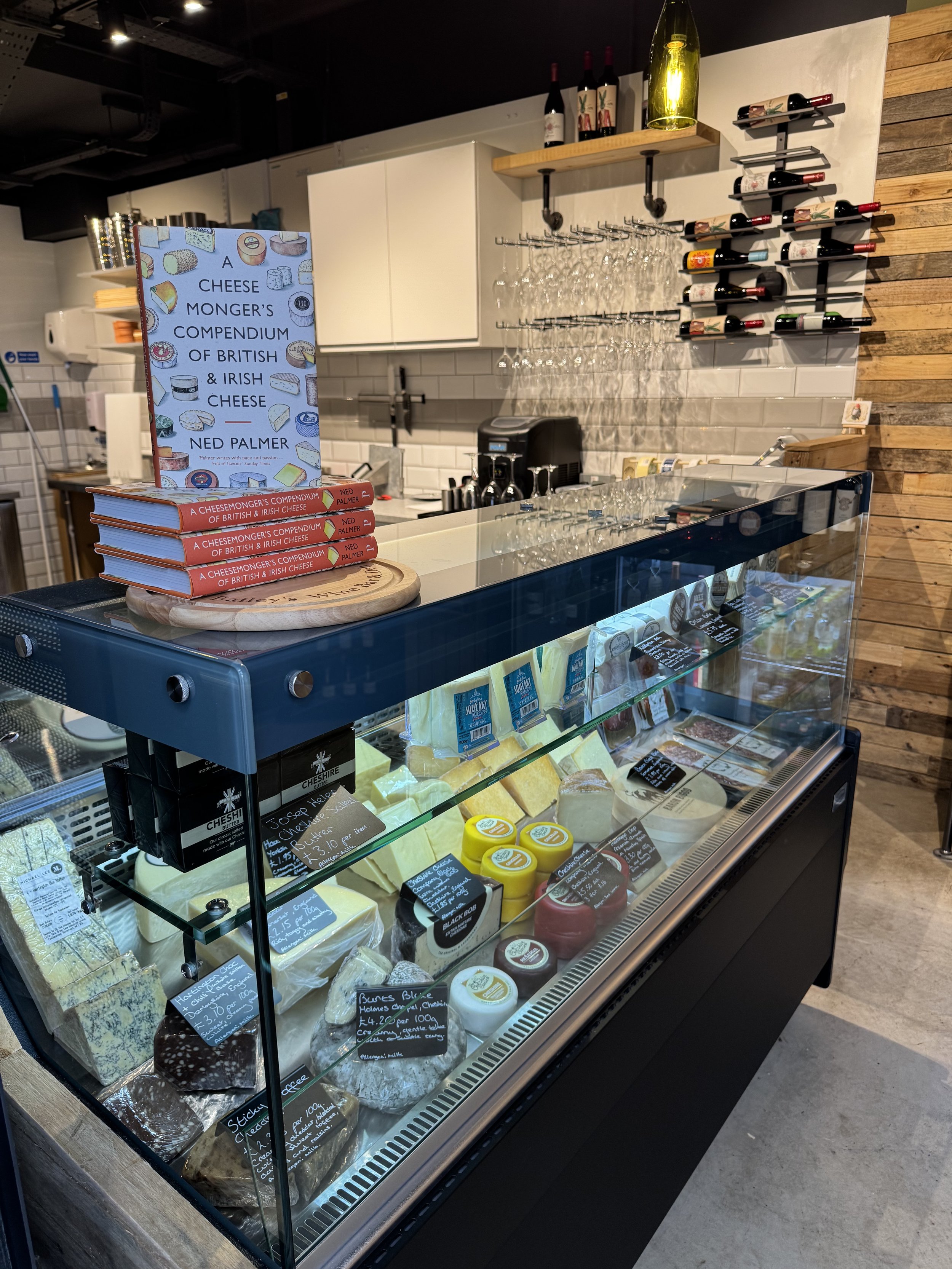 Cheese display in a shop with an assortment of cheeses, books stacked on a counter, wine bottles, and hanging glasses in the background.