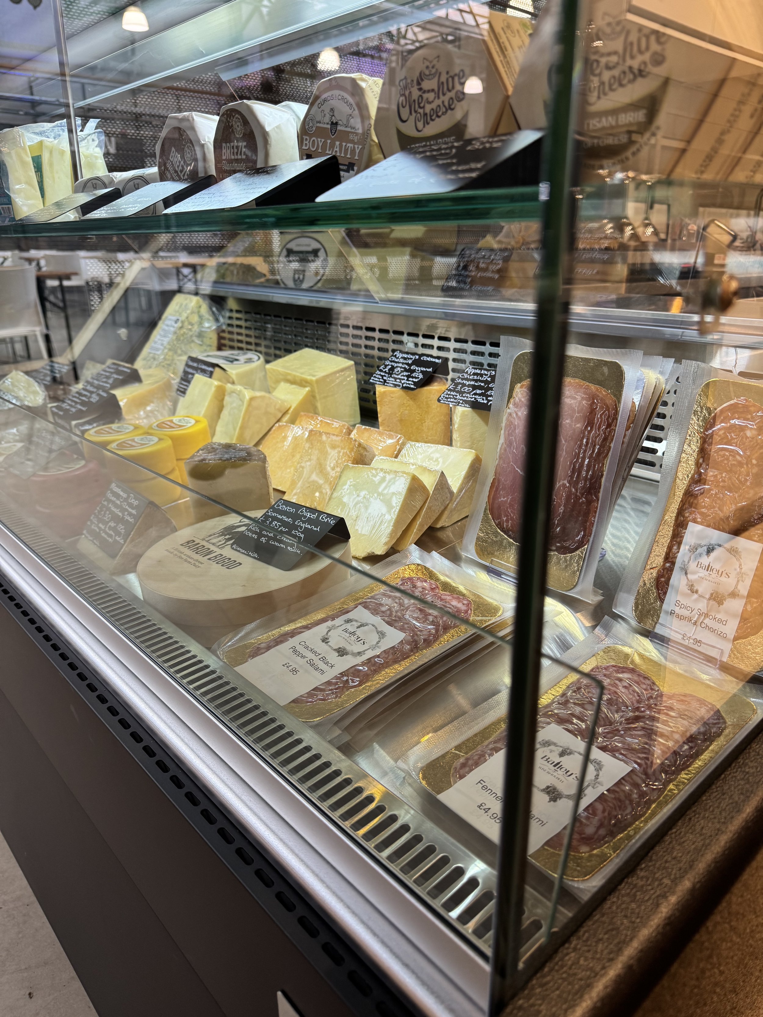 A deli display case with assorted cheeses and cured meats. Various cheese types are stacked, some labeled with signs. Packaged cured meats are arranged at the bottom. Soft lighting and glass reflection are visible.