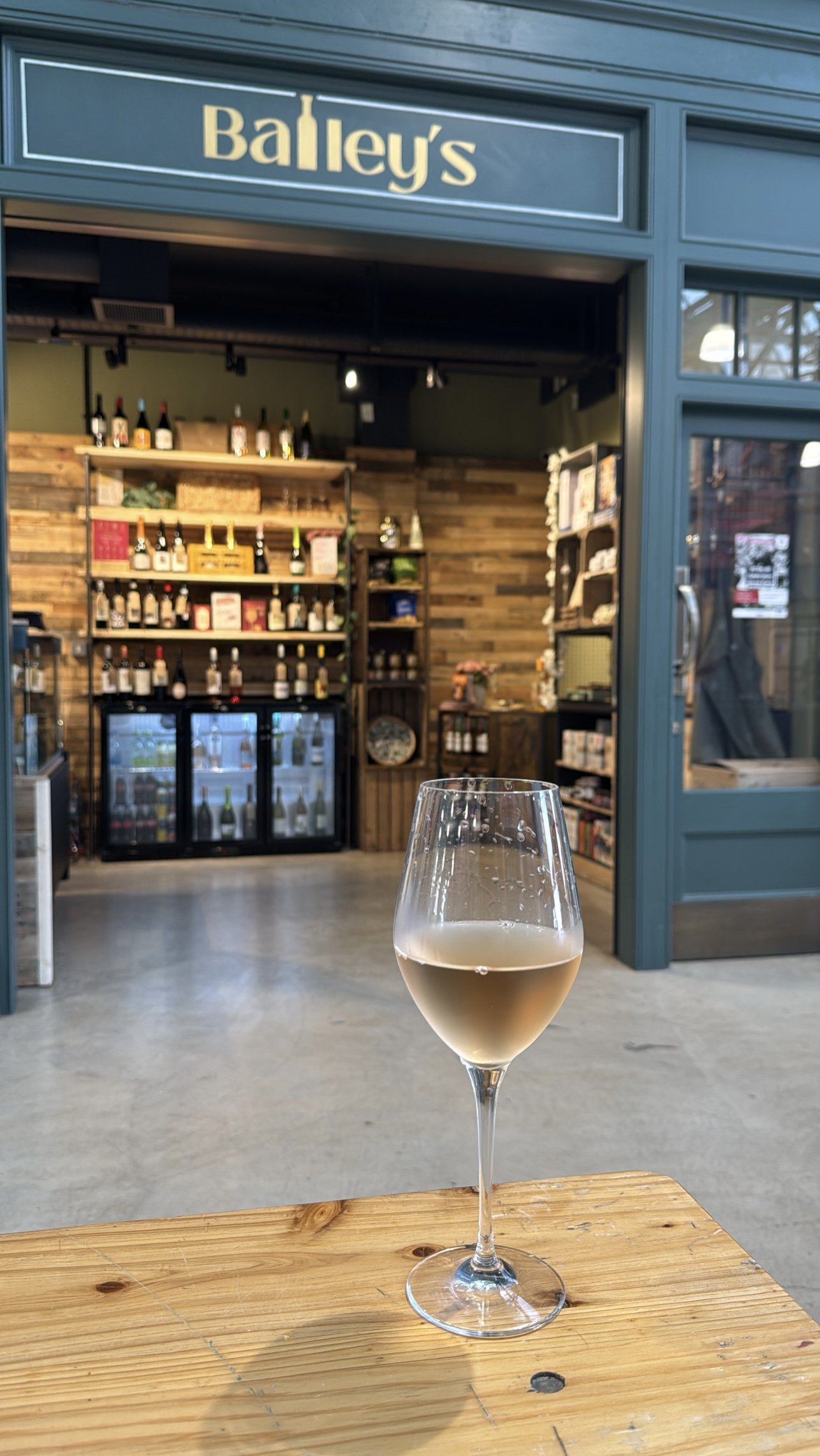 A wine glass with a light-colored drink on a wooden table in front of a store named Bailey's. Inside the store, shelves display various bottles and products.
