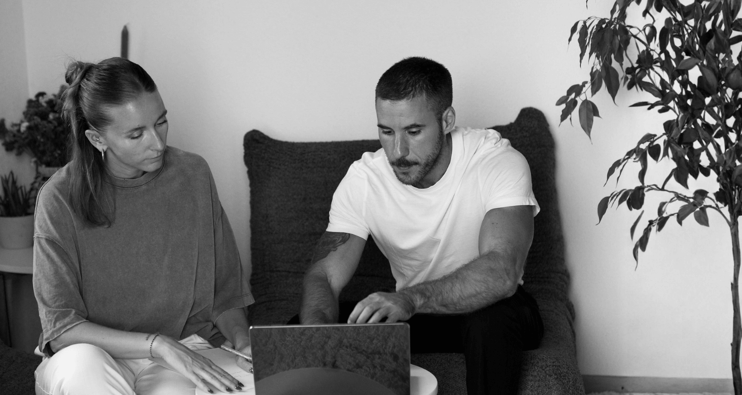 Deux personnes, un homme et une femme, regardent un ordinateur portable dans un salon, assis sur un canapé. Un arbre d'intérieur est visible à droite, et une plante est sur une table à gauche.