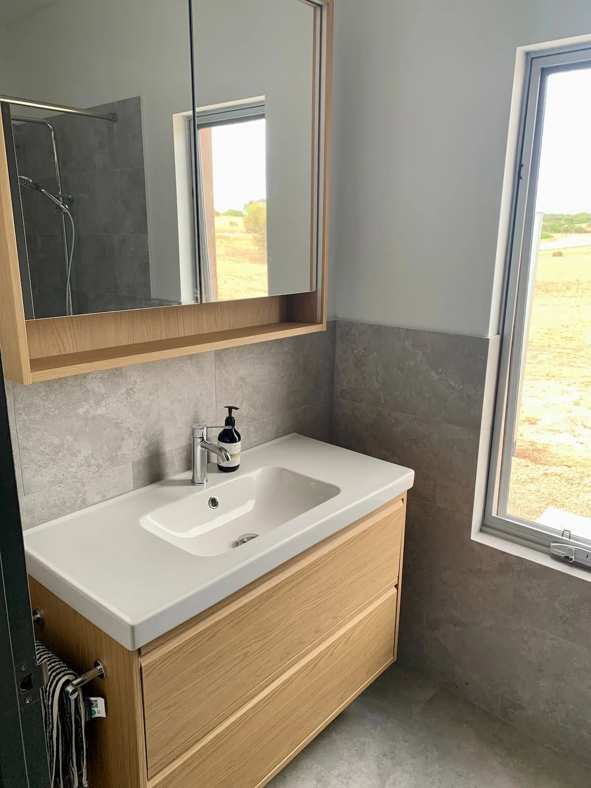 Modern bathroom with a wooden vanity, white sink, black soap dispenser, gray tiled wall, large mirror, and window showing an outdoor landscape.
