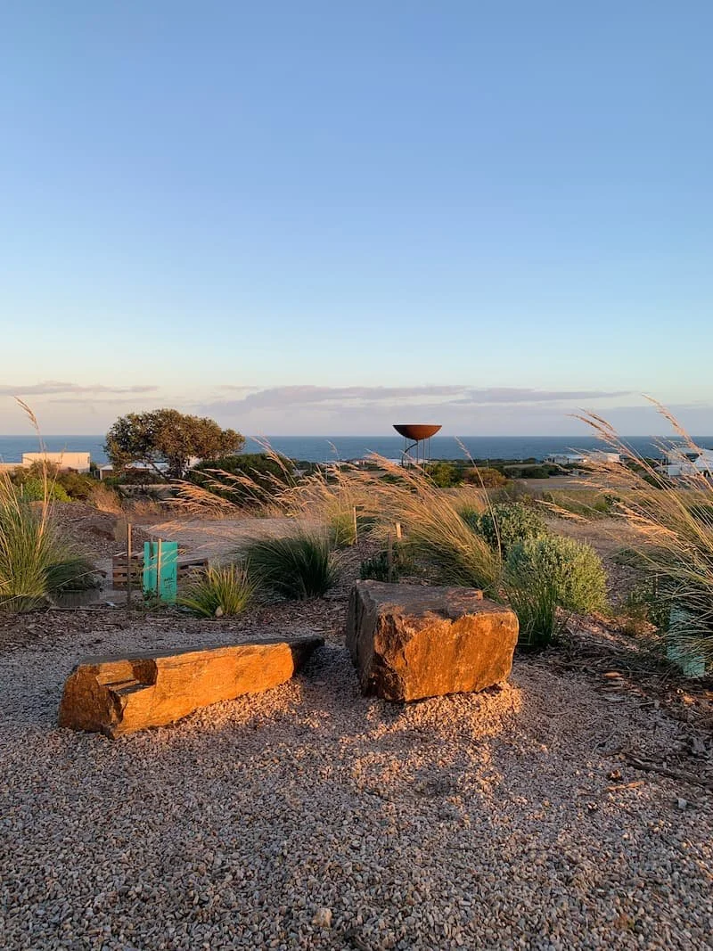 A scenic landscape with rocks, prairie grass, trees, and a view of the ocean under a clear sky.