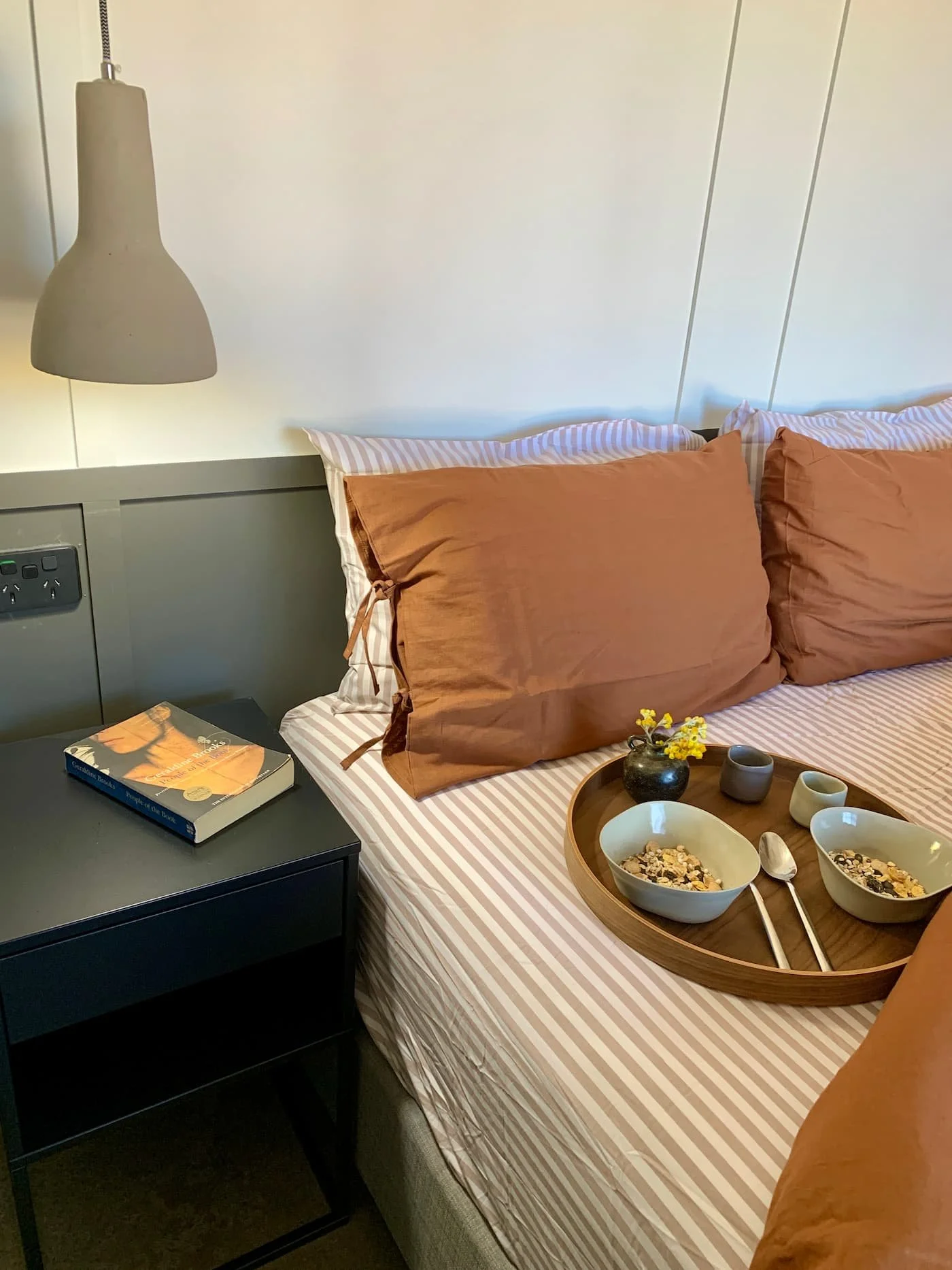 A neatly made bed with two large rust-colored pillows on a striped bedsheet. There is a wooden tray on the bed with bowls of cereal, small vases, and a spoon. A black nightstand next to the bed has a book and a dark-colored lamp hanging above.