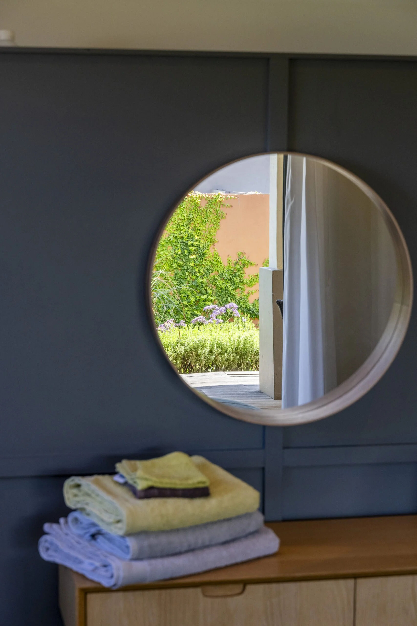Stacked folded towels on a wooden surface with a view of a garden through a circular opening in a dark blue panel.