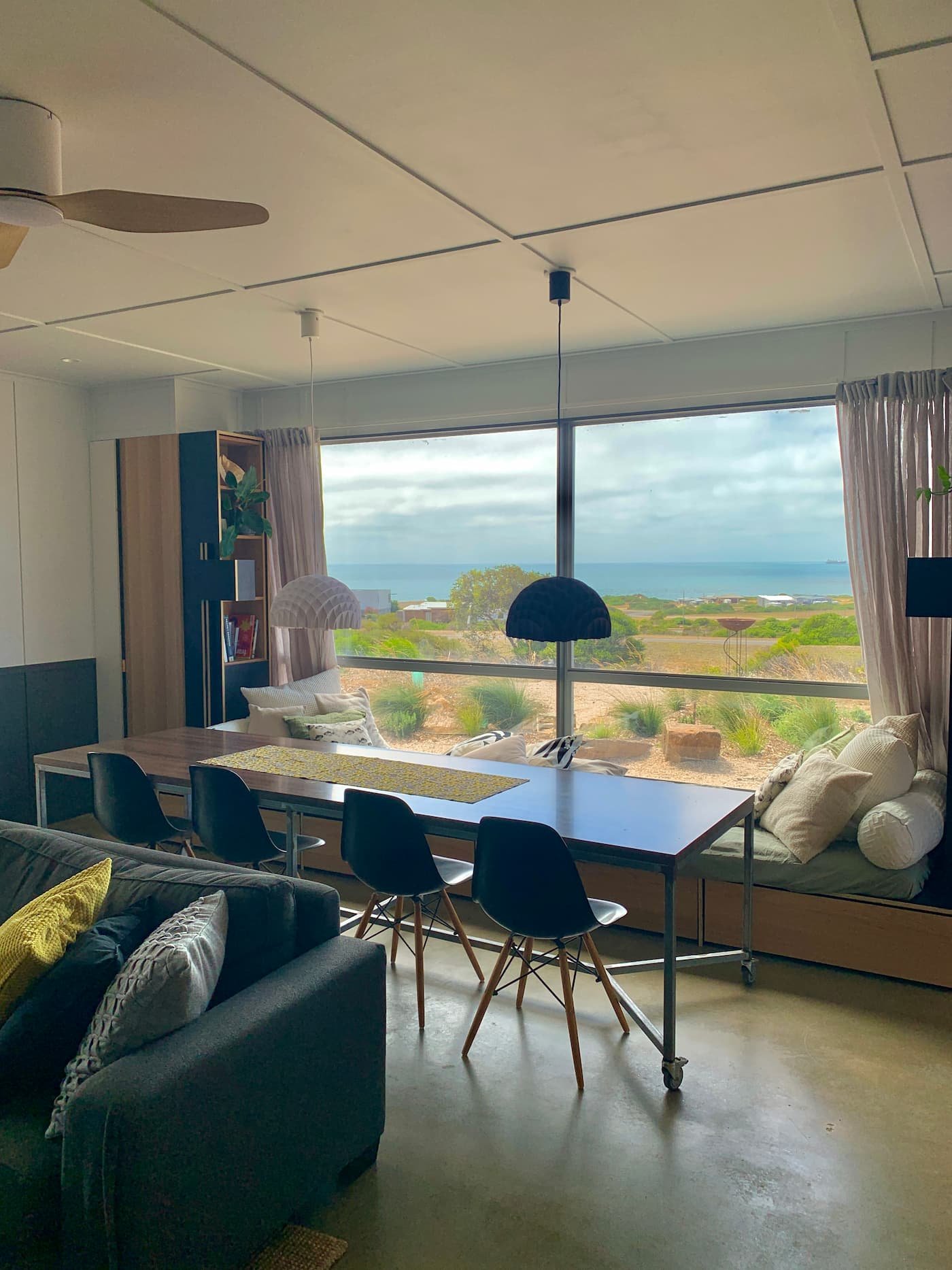 Living room with large window view of the ocean, featuring a dining table with four black chairs, a gray sofa with yellow and gray pillows, window seat with cushions, and a bookshelf. Pendant lights hang above the table.