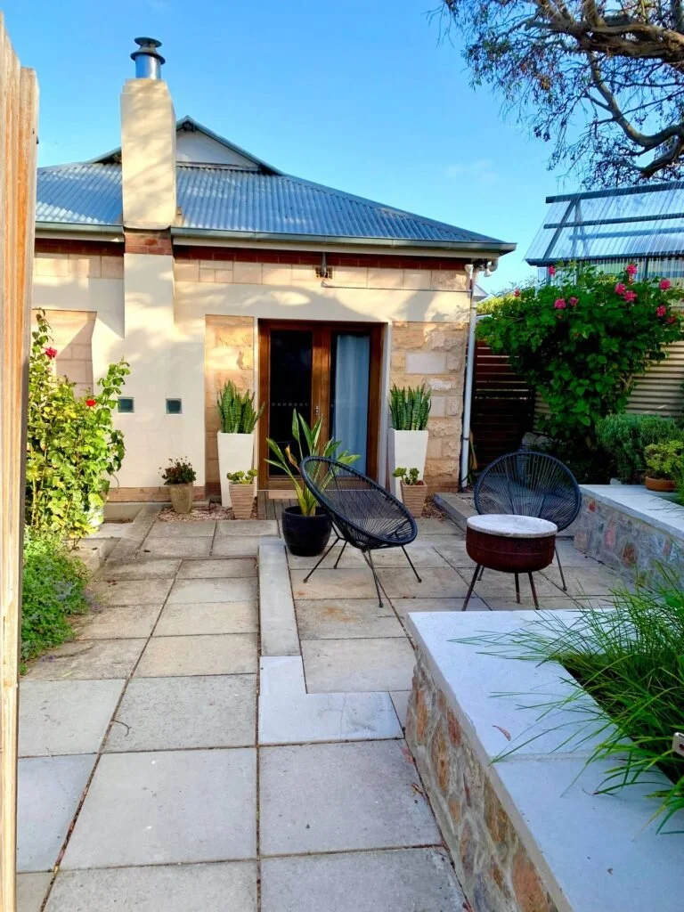 A small outdoor patio area with black circular chairs, a round side table, tall potted plants, and a house with a metal roof and brick walls. There is a glass door leading into the house, and greenery on both sides of the patio.