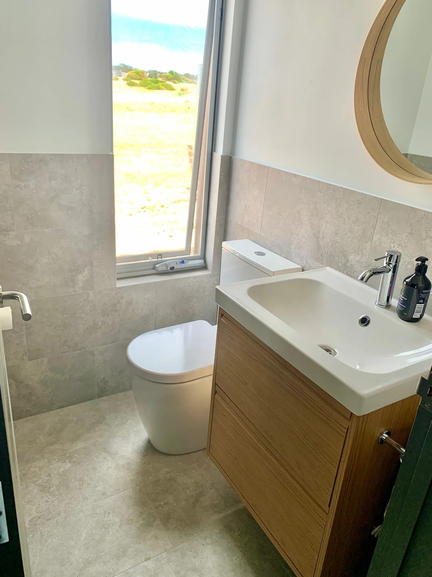Modern bathroom with a white toilet, a small white sink with a wooden cabinet, a round mirror, and a window showing an outdoor landscape.