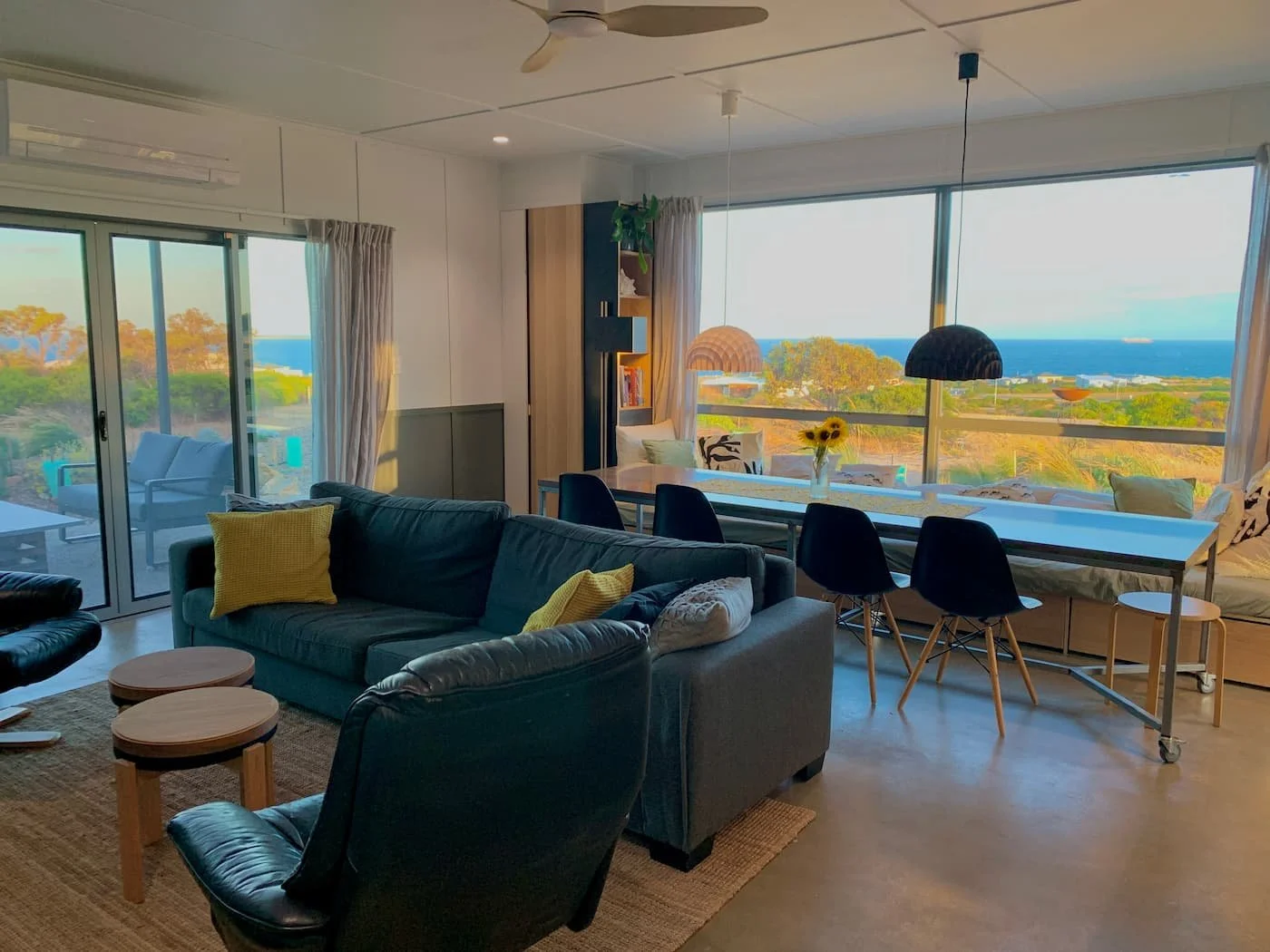 Living room with large windows showing a view of trees, ocean, and sky, furnished with sofas, chairs, a dining table, pendant lights, and decorative pillows.