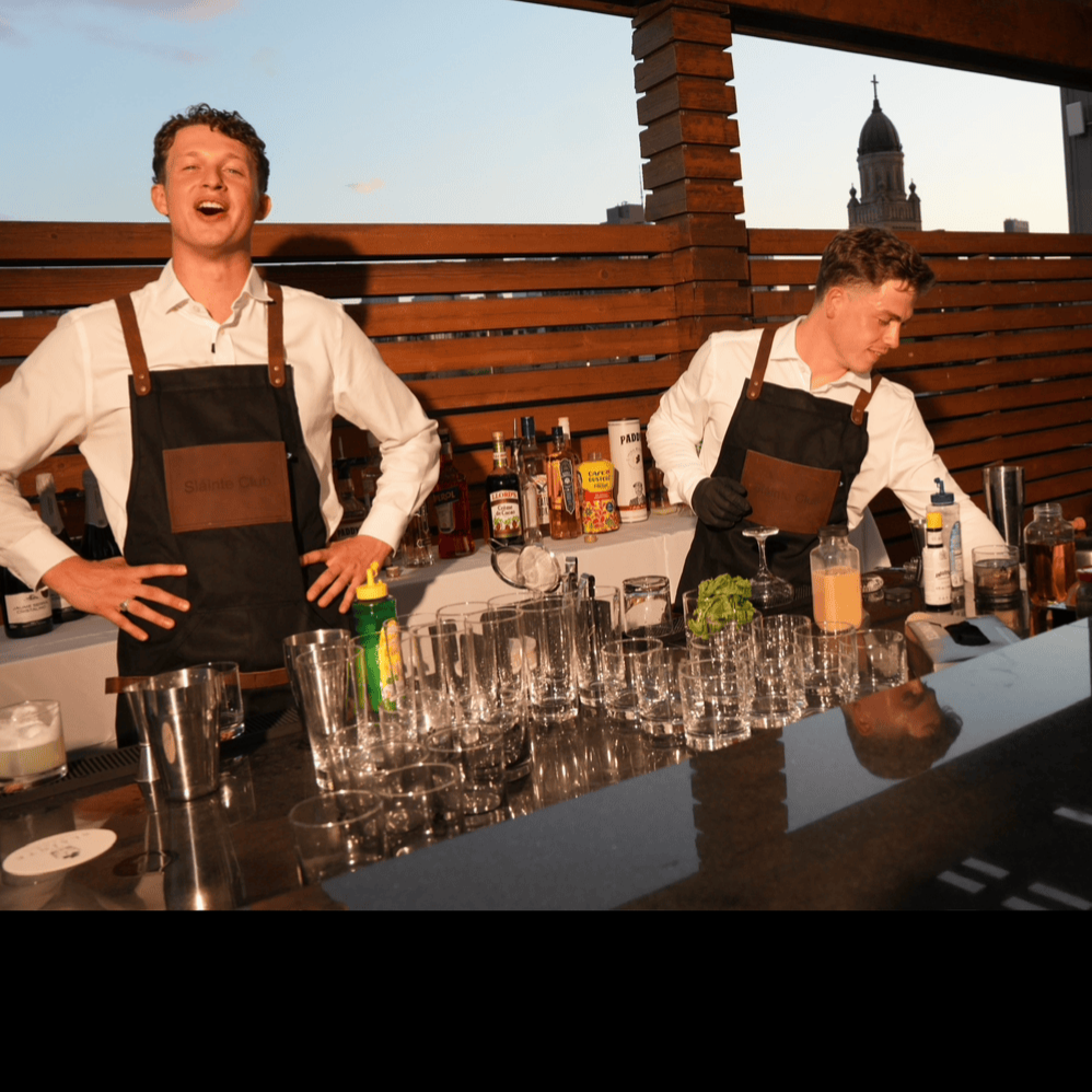Two male bartenders in white shirts and black aprons preparing drinks on a rooftop bar at sunset, with a city skyline in the background.