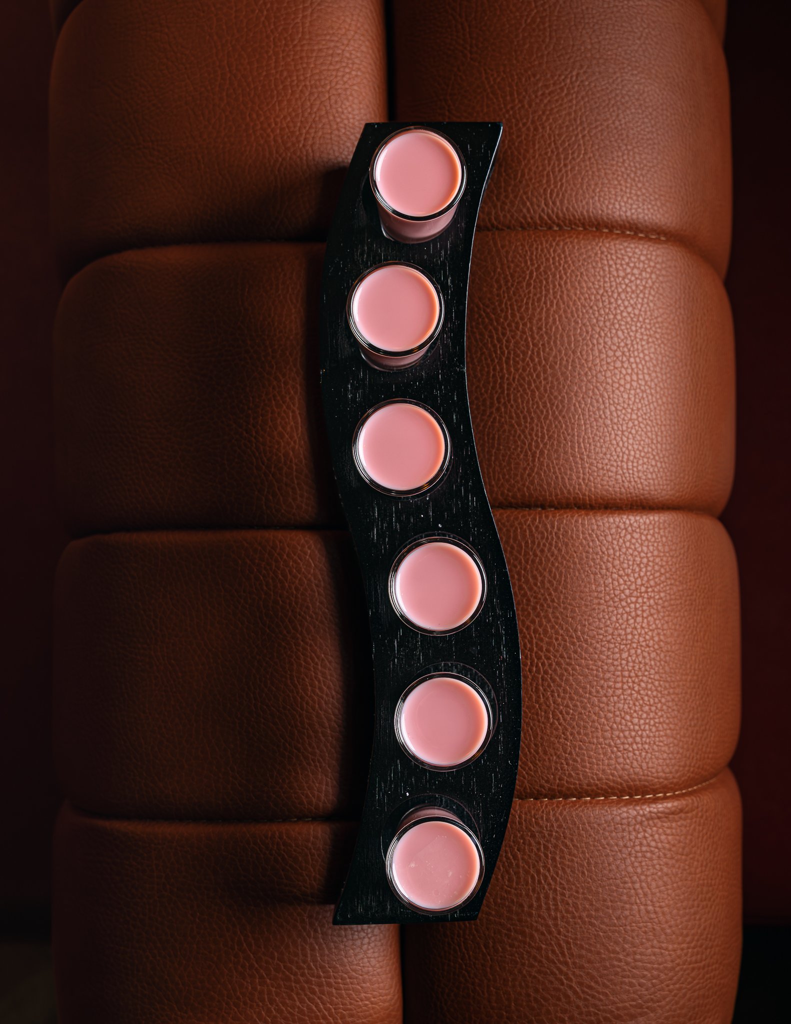 Top view of four glasses of light pink beverage on a black tray, placed on brown leather upholstery.