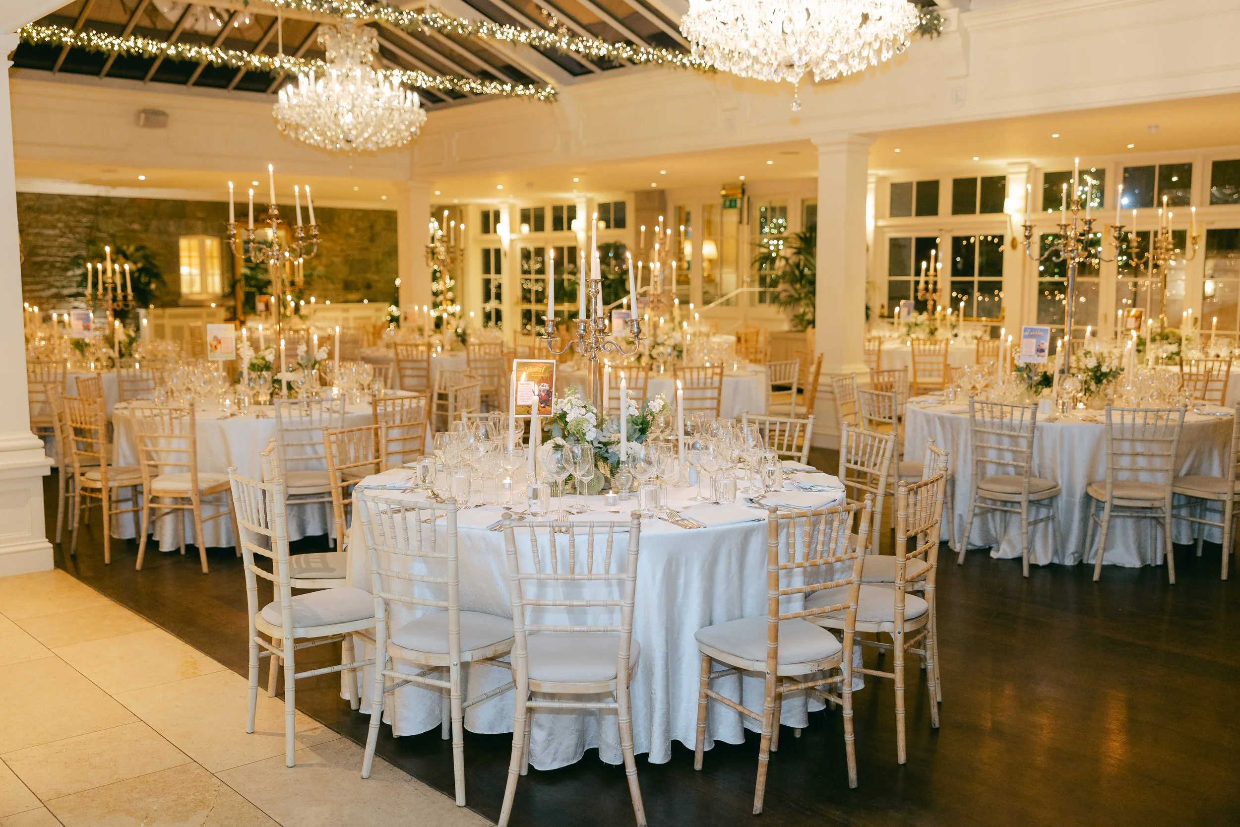 Elegant banquet hall decorated for a formal event with white tablecloths, floral centerpieces, tall candelabras with candles, and string lights and chandeliers hanging from the ceiling.