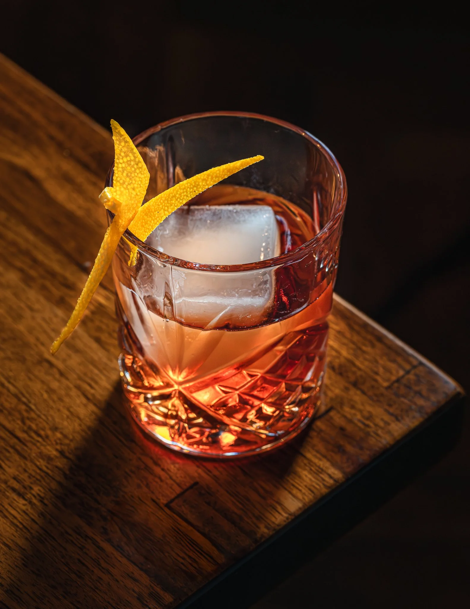A pinkish-red glass tumbler with a textured pattern, filled with a clear beverage with ice cube, garnished with two curled lemon peel twists, on a dark wooden table.