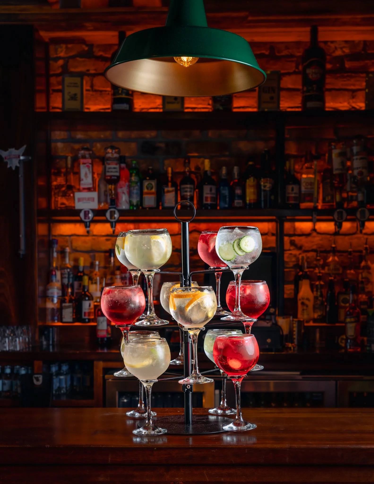 A layered display of colorful cocktails in large wine glasses on a bar counter, with a dimly lit bar and liquor bottles in the background.