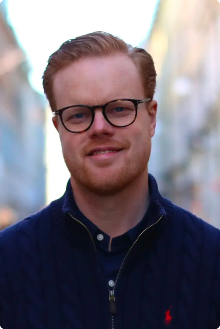 Portrait of a young man with red hair and glasses, smiling, wearing a navy blue jacket, outdoors in a city setting.