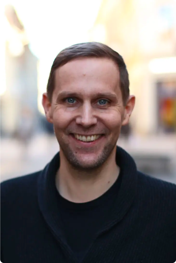 Close-up of a smiling man with short brown hair, blue eyes, wearing a black shirt, outdoors in an urban setting with blurred buildings in the background.
